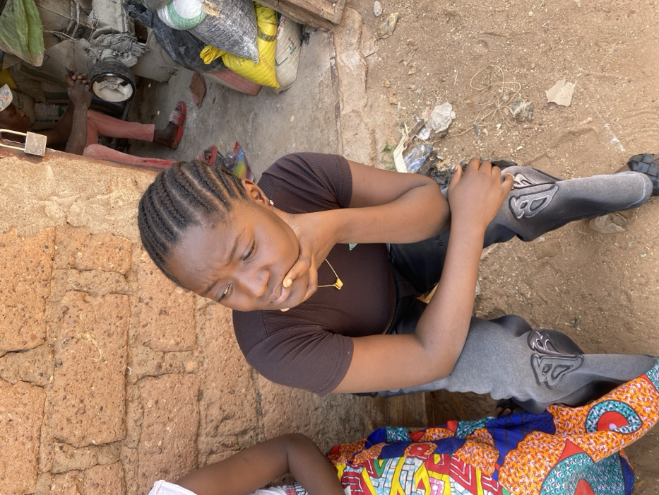 The scene is set outdoors in Rido, Nigeria, against a reddish-brown mud brick wall. In the foreground, a young Black female is seated, facing left, with her chin resting on her right hand. She has dark braided hair, wears a dark brown ribbed short-sleeved top, grey sweatpants with a prominent 'B' logo on the knees, a gold-colored necklace, and black woven slide sandals. To her left, another person is partially visible, wearing a colorful, patterned fabric. To the right, through a doorway, a male figure wearing a cap, light-colored pants, and red slide sandals operates a piece of machinery, possibly a grinding machine. Black and yellow sacks are stacked nearby, and a brass padlock hangs from the doorframe. The ground is unpaved reddish-brown earth with scattered debris.