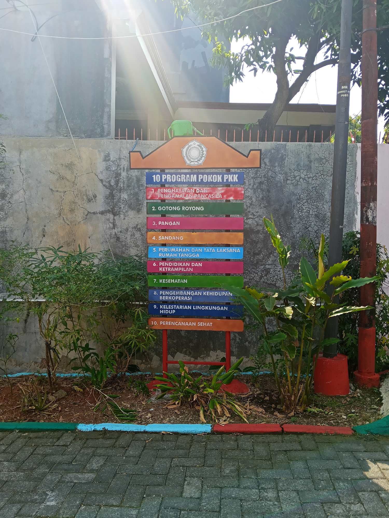 The image shows a sign detailing the "10 PROGRAM POKOK PKK" (10 Main PKK Programs). The sign is multicolored with each program listed on a separate colored panel. The PKK is a national family welfare movement in Indonesia. The programs listed are: 1. Penghayatan dan Pengamalan Pancasila (Appreciation and Practice of Pancasila), 2. Gotong Royong (Mutual Cooperation), 3. Pangan (Food), 4. Sandang (Clothing), 5. Perumahan dan Tata Laksana Rumah Tangga (Housing and Household Management), 6. Pendidikan dan Keterampilan (Education and Skills), 7. Kesehatan (Health), 8. Pengembangan Kehidupan Berkoperasi (Development of Cooperative Life), 9. Kelestarian Lingkungan Hidup (Environmental Sustainability), and 10. Perencanaan Sehat (Healthy Planning).

The sign is mounted on a weathered concrete wall. To the left and right of the sign are various plants, with dry leaves and soil on the ground. The area in front of the wall is paved with interlocking gray bricks. In the background, there is a building with a white facade and a tiled roof. Sunlight streams in from the left, casting strong shadows and creating lens flare effects. A dark pole, likely a utility pole, is visible to the right of the sign, with some writing on it. The overall scene suggests an outdoor communal area or educational facility.

While the image captures elements of daily life and community programs in Indonesia, there is no specific information in the visual content to pinpoint the exact location within Semarang. The scene is generic enough to be found in many urban or suburban areas across Indonesia.