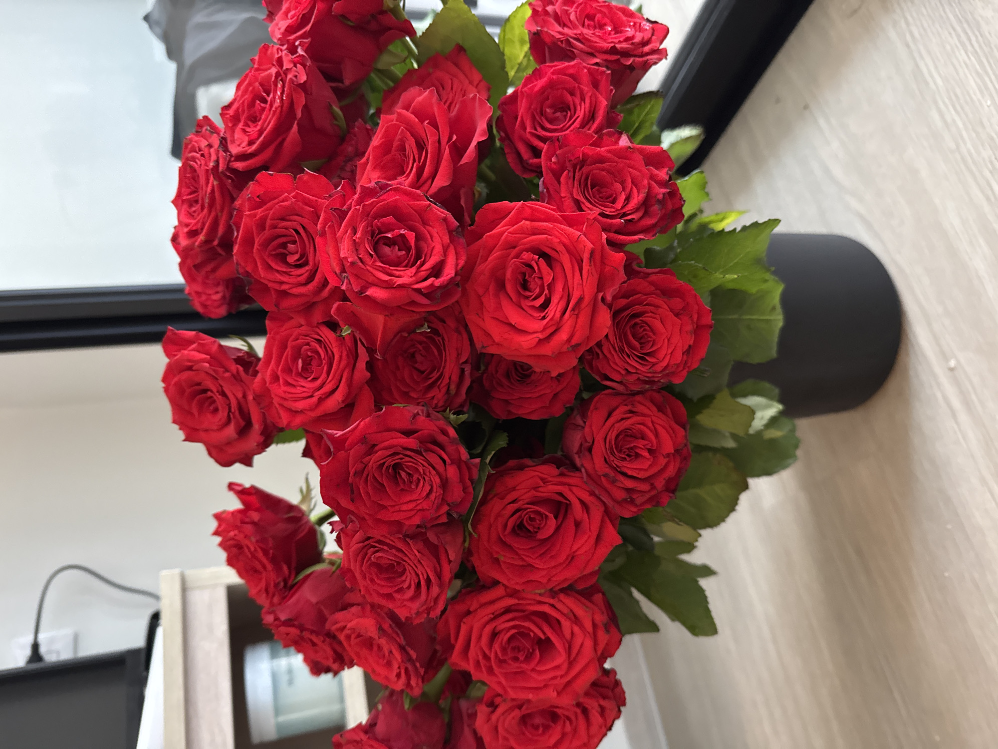 A bouquet of red roses is displayed in a black vase on a wooden surface. The roses are a deep red color and are densely packed together, with green leaves visible among them. The background shows a blurred indoor environment with a shelf and a portion of a window. There is no indication of Kathu, Thailand in the image.
