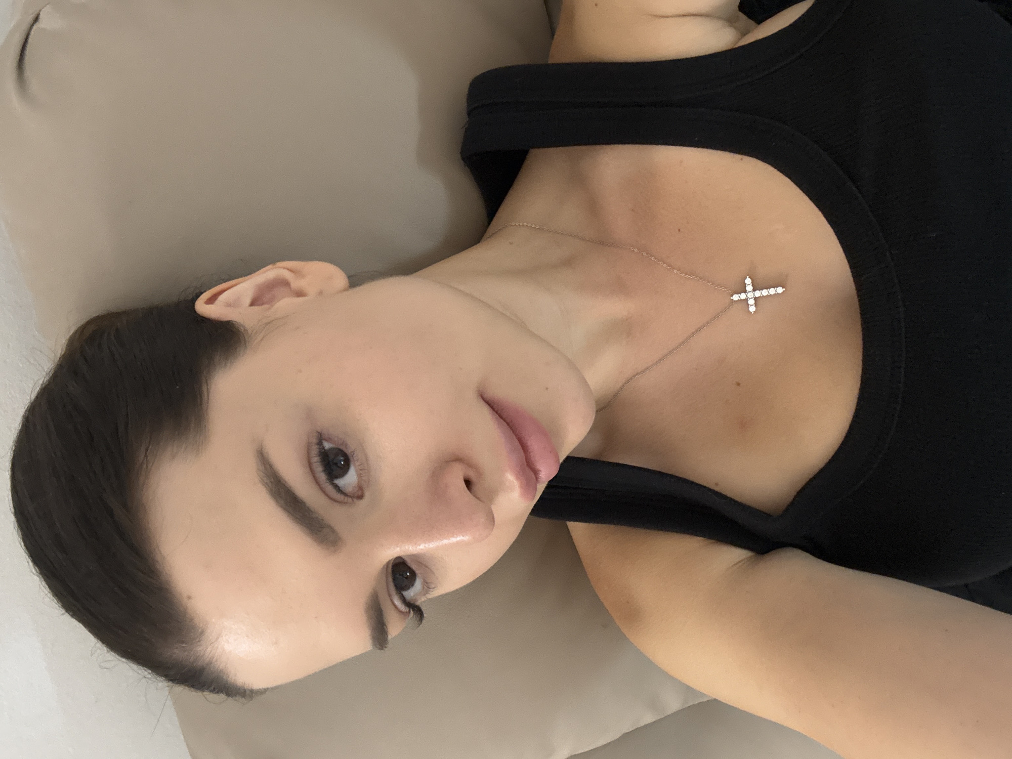 A person is lying down on a light brown surface, possibly a couch or bed. They are wearing a black tank top and a delicate silver necklace with a cross pendant. The cross is adorned with small, clear stones, likely diamonds. The person's hair is dark and pulled back, with some strands framing their face. Their face is turned towards the camera, with their left eye visible. They have full lips and dark eyebrows. The lighting suggests it might be indoors, with soft lighting. The orientation of the image is a rotation of 90 degrees counterclockwise from a standard upright view. The background is a neutral tone, creating a simple and focused composition on the subject. No visible text is present in the image. The context of Prague, Czechia does not provide any specific visual cues within the image itself.