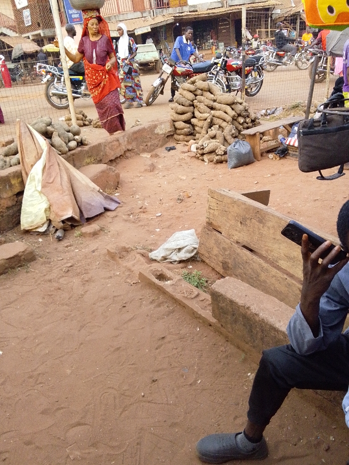 A bustling outdoor market features a woman carrying goods on her head. Piles of yams sit near a makeshift bench, where a man in a blue shirt and dark pants uses a cell phone. Motorcycles line the street behind the vendor, with other individuals walking in the background. The ground is reddish-brown dirt, and buildings are visible in the distance.