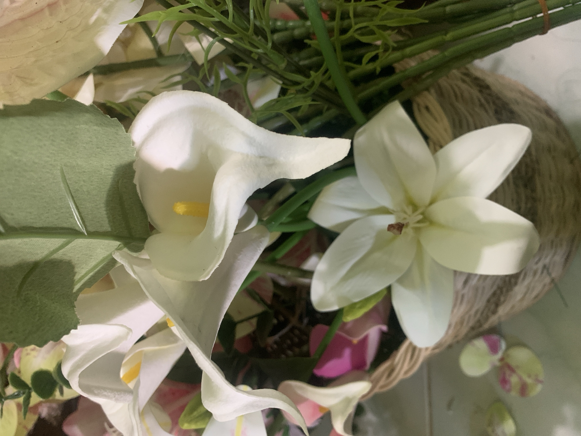 A close-up, eye-level, still life photograph captures a floral arrangement in a woven basket. The basket, made of light brown fibrous material, is positioned slightly off-center to the right. A prominent white calla lily with a bright yellow stamen is the focal point, positioned in the upper half of the frame, with its curved spathe facing downwards. To the lower left of the calla lily, another white calla lily is partially visible. In the foreground, a white flower with five pointed petals and a light brown center rests on the edge of the basket. Behind the calla lilies, various green leaves and stems are visible, including a large, textured green leaf in the upper right quadrant. There are also hints of pink and lighter colored flowers in the background. The lighting appears to be natural, casting soft shadows. The overall impression is a close-up view of an artificial floral display. The context of Kuala Pembuang, Indonesia, is not evident in the visual elements of the photograph itself.