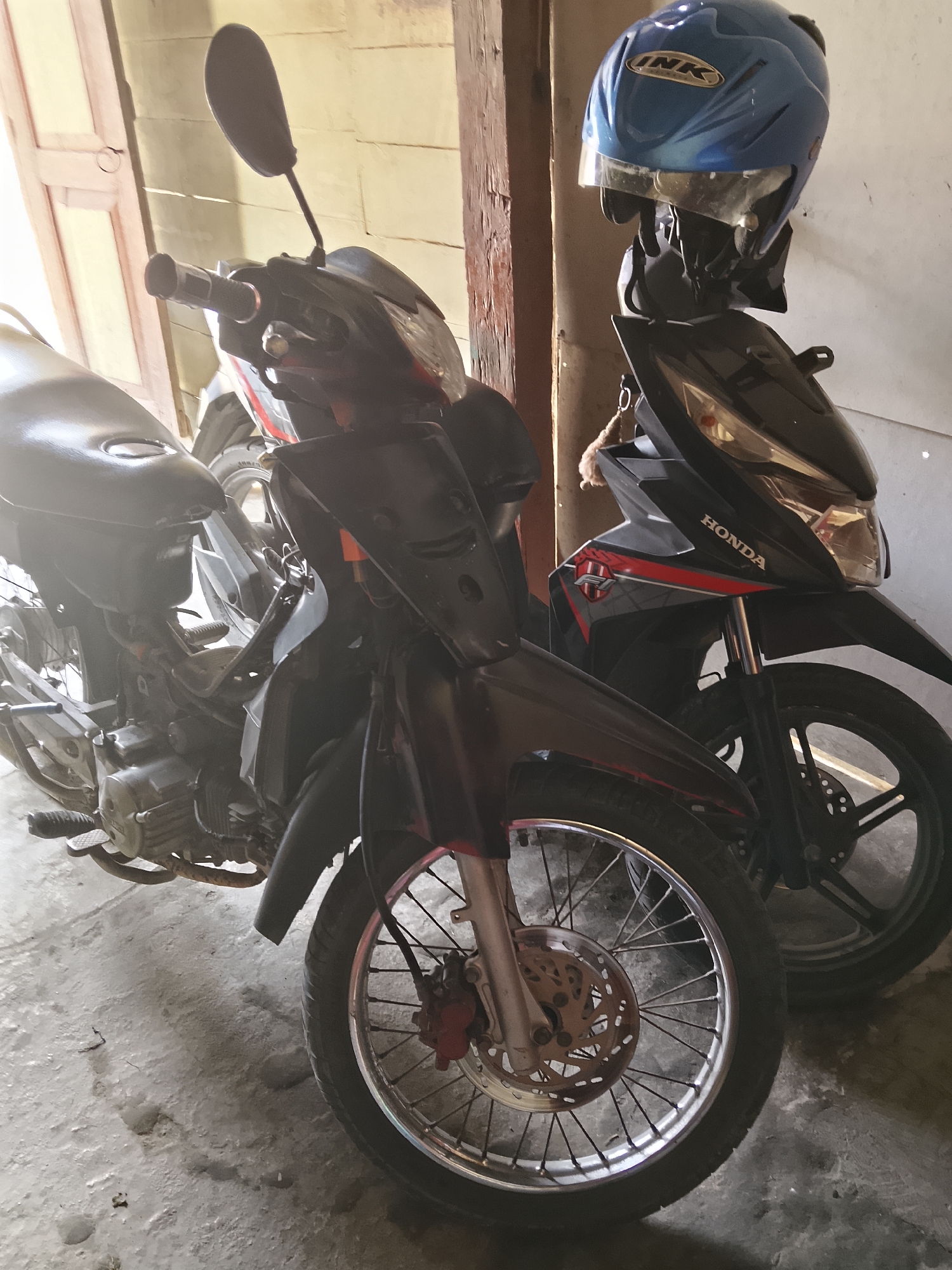 Two motorcycles are parked next to each other in a dimly lit indoor space, possibly a garage or carport, with natural light filtering in from an open doorway on the left. The motorcycle in the foreground on the left is black and appears to be a semi-automatic scooter with a large seat and exposed engine components. The motorcycle on the right is also black and red and is a Honda, with a blue helmet hanging from its handlebars. The ground is concrete and appears to be wet in some areas, with visible footprints and debris. The background shows a textured wall and wooden support beams. The scene suggests a residential or utilitarian setting in Kasiman, Indonesia.