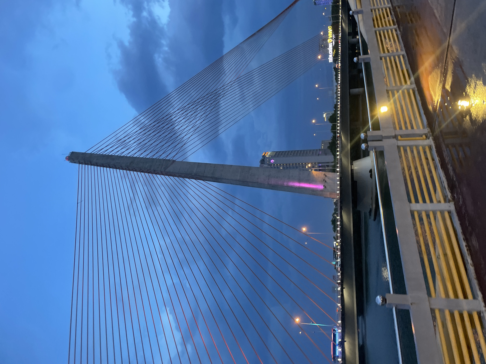 The image depicts a cable-stayed bridge at dusk, with a dramatic sky filled with dark clouds. The bridge's concrete pylon and numerous red cables are prominent subjects, stretching across the frame. In the background, city lights twinkle, and a tall building with pink and purple lights is visible. A yellow-railed pedestrian walkway runs along the right side of the bridge, with wet pavement suggesting recent rain. Streetlights illuminate the walkway, casting reflections on the wet surface. The overall mood is serene and slightly melancholic, enhanced by the twilight setting and the vast expanse of the sky. Visible text on a building in the background reads "TIỀU CƯỜNG VNVC". The time of day is clearly evening or dusk, with the sky transitioning from day to night. The location is identified as Đà Nẵng, Vietnam. No people are clearly visible in the image.