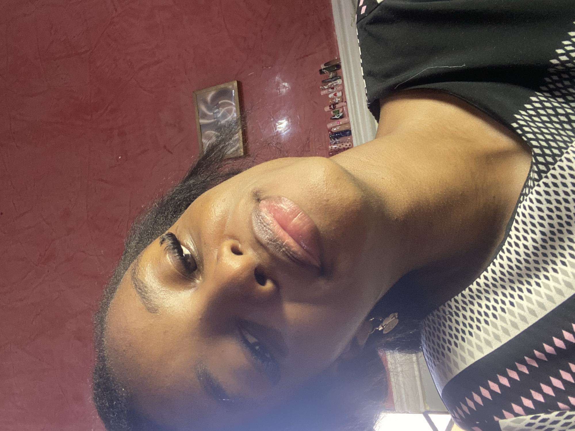 The image shows a close-up, low-angle selfie of a young Black woman indoors. She has dark skin, dark hair, and is wearing false eyelashes and lip gloss. She is wearing a black shirt with a geometric pattern. In the background, a red textured wall is visible, with a small framed picture hanging on it. Below the picture, a row of what appear to be artificial nails are displayed on a shelf or ledge. The lighting suggests an indoor setting, possibly a bedroom or vanity area. The image does not provide any specific visual cues to pinpoint a location within Jos, Nigeria.