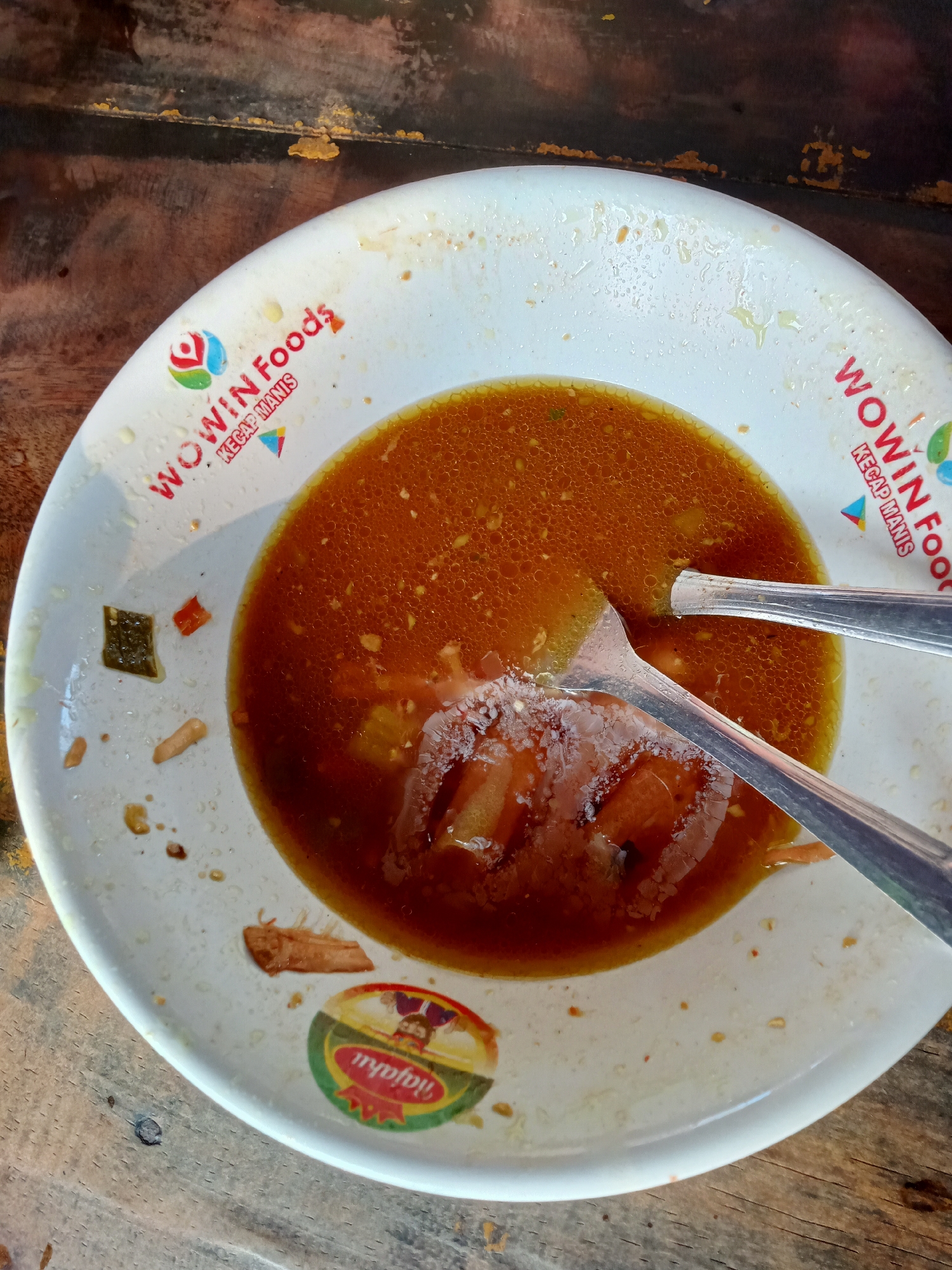 A close-up, overhead shot shows a nearly empty white bowl with residue of brown broth and some solid food pieces on a wooden surface. The bowl has red text that reads "WOWIN FOODS KECAP MANIS" and a circular sticker with a cartoon character that appears to say "Tijayu". Two silver spoons are resting in the bowl. The wooden surface is a table, possibly in a restaurant. The lighting is natural, coming from above. The image is sharp and clear. The location of Majan, Indonesia is not visible in the image.