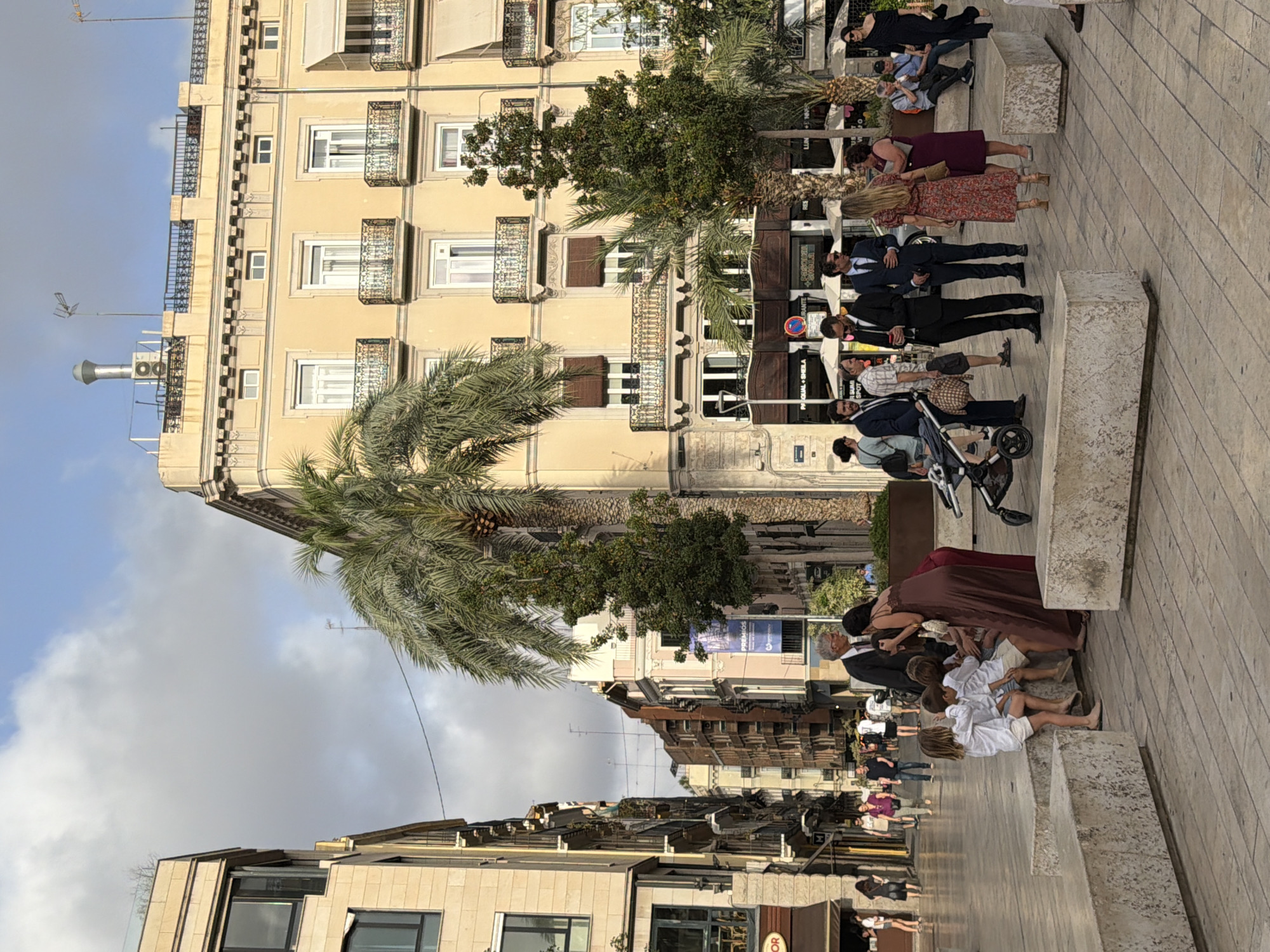An eye-level, full shot captures a street scene in an urban environment, likely Europe, given the architecture. The day is bright, with the sun casting subtle shadows. A large, multi-story building with a beige facade and ornate balconies dominates the right side of the frame. The building features numerous windows, some with shutters, and decorative cornices. A prominent palm tree stands in front of the building, with its fronds reaching towards the sky.
In the foreground, a group of people are gathered on a paved plaza. Three young children, dressed in white shirts and shorts, are seated on a large concrete block. An adult woman, wearing a long, dark brown dress, stands over them, appearing to interact with one of the children. Next to her, another woman in a maroon dress stands with her arm extended.
Further back, a group of men in suits are standing and talking. One man is looking at his phone, while another appears to be gesturing. A baby stroller is parked nearby. In the background, other pedestrians walk along the street, and more buildings line the sides of the street. The overall impression is one of a lively public space.