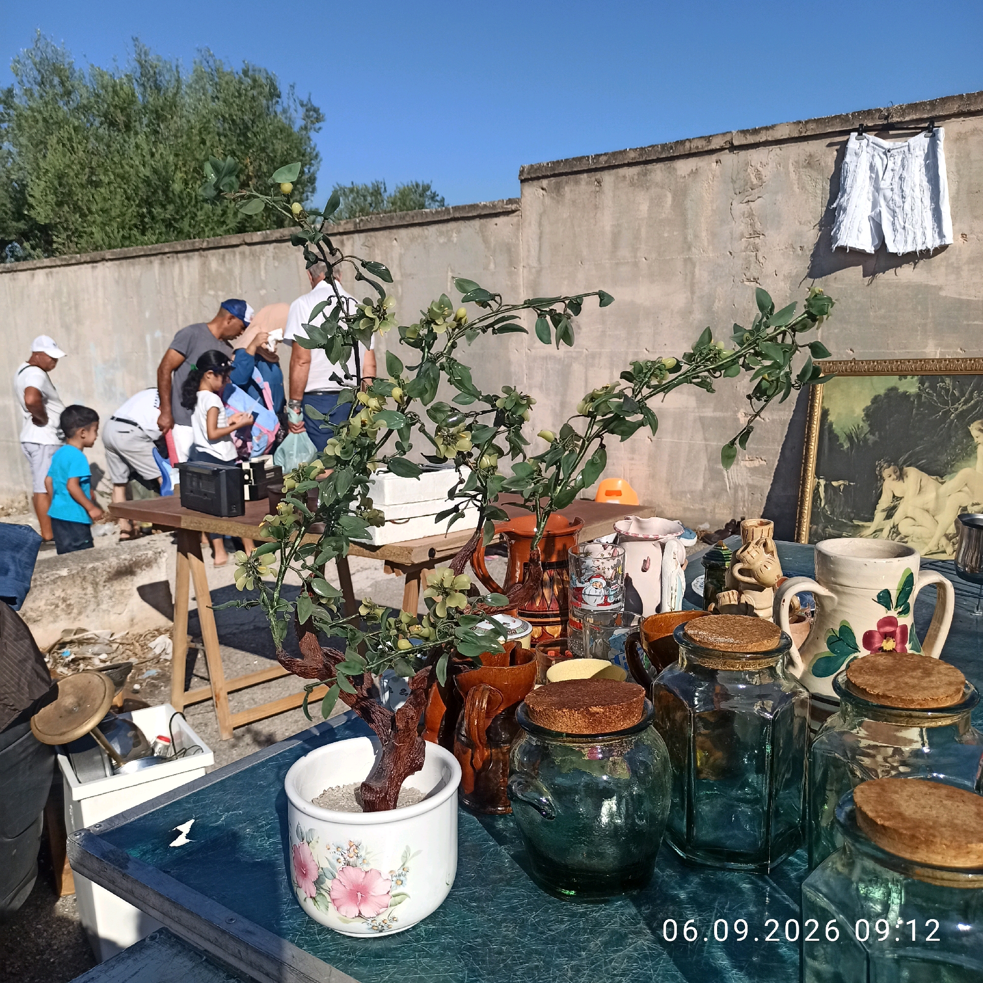 The image depicts an outdoor flea market scene, likely in Sannicandro di Bari, Italy, given the provided context. In the foreground, a table is laden with various items, including glass jars with cork stoppers, ceramic pitchers, and decorative plant pots with artificial greenery. A framed painting of nudes is visible in the background, leaning against a weathered concrete wall. Several individuals, including adults and children, are browsing the merchandise. Some are engaged in conversation, while others examine items on display. A pile of discarded items and a white bin are visible to the left of the table. The setting appears to be a sunny day, with a clear blue sky. A timestamp "06.09.2026 09:12" is overlaid on the image.