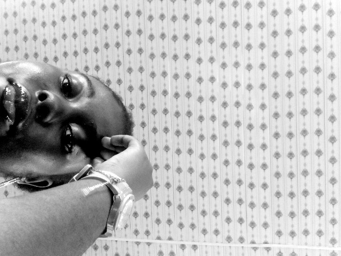 This black and white image, oriented ninety degrees clockwise, depicts a single individual indoors. The subject, appearing to be an adult with dark skin, is positioned in the lower left of the frame (when viewed with correct orientation), with their head tilted. Their lips are prominent and glossy, and their eyes are directed towards the viewer. A metallic wristwatch with a chain attachment is visible on the subject's left wrist. Their left hand is raised, with fingers near the side of the head. The background consists of a wall covered in patterned wallpaper, featuring repeating vertical rows of light-colored rhomboid or floral motifs against a darker striped background. The specific location within Ejule, Nigeria, is not discernible from the visual evidence, but the scene appears to be a residential or indoor setting.