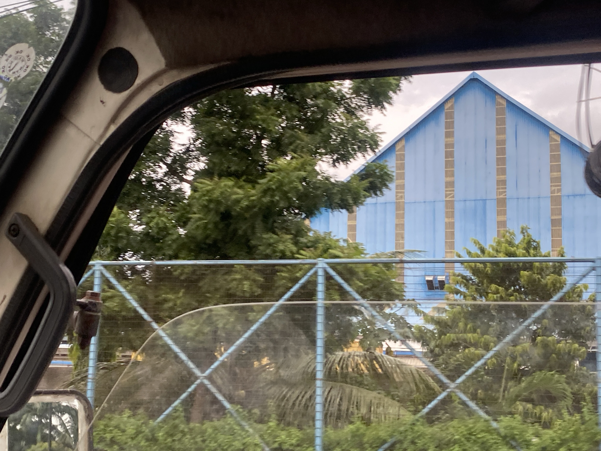 The image, taken from inside a vehicle in Lagos, Nigeria, captures a view through the windshield and side window. Dominating the background is a large, light blue corrugated metal building with a prominent pitched roof. Vertical, light brown or beige panels are spaced along the facade of the building. In front of the building, a dense canopy of green trees obscures the lower portion of the structure.
A light blue metal fence with a diamond-shaped mesh pattern runs horizontally across the mid-ground of the image. This fence is positioned in front of the trees and the building. Within the fence, more greenery and foliage are visible, including what appears to be palm fronds.
In the foreground, the interior of the vehicle is visible on the left side. This includes the A-pillar of the car, a black circular object (possibly a speaker or a sensor), and a portion of the side mirror. There are also round stickers with markings on the A-pillar, one of which has "OK" visible. The windshield shows reflections of trees.
The overall lighting suggests an overcast or slightly hazy day. No people are discernible in the image. The scene depicts an outdoor environment, likely in an industrial or semi-industrial area of Lagos, given the large building and fencing.
