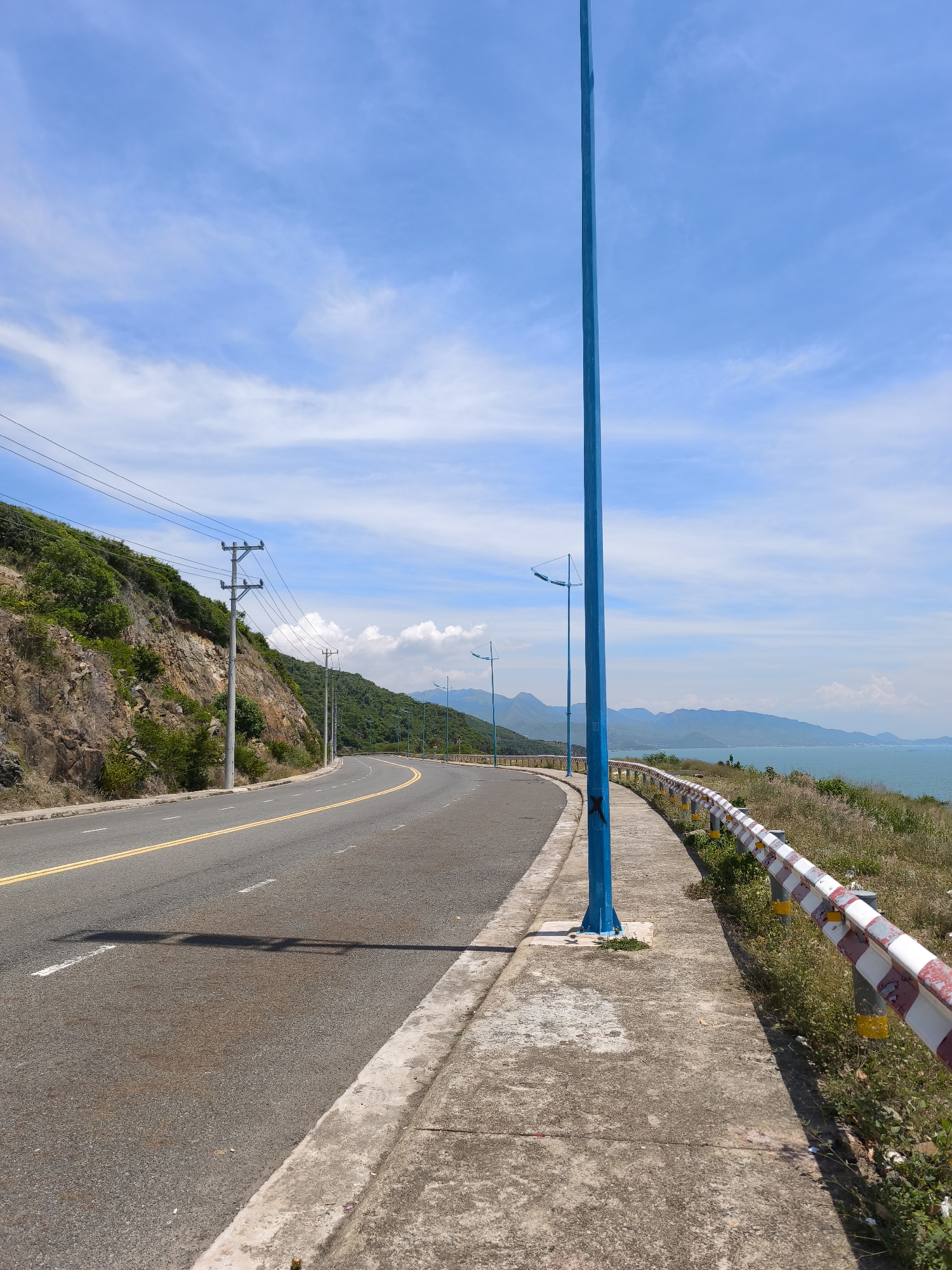 The image shows a coastal road curving to the left. The road is asphalt with a double yellow line down the center and white dashed lines indicating lanes. On the left side of the road is a steep, rocky hillside covered in green vegetation. Utility poles with wires run along the hillside. On the right side of the road is a concrete sidewalk, a guardrail with red and white stripes, and dry grass. A tall blue pole with a light fixture stands on the sidewalk. In the background, there is a body of water, likely the sea, with a mountainous coastline and a hazy blue sky with white clouds. The image is taken from the perspective of someone standing on the sidewalk, facing the curve of the road. The lighting suggests it is daytime, with the sun casting shadows. There are no people visible in the image. The location is identified as Bac Nha Trang Ward, Vietnam.