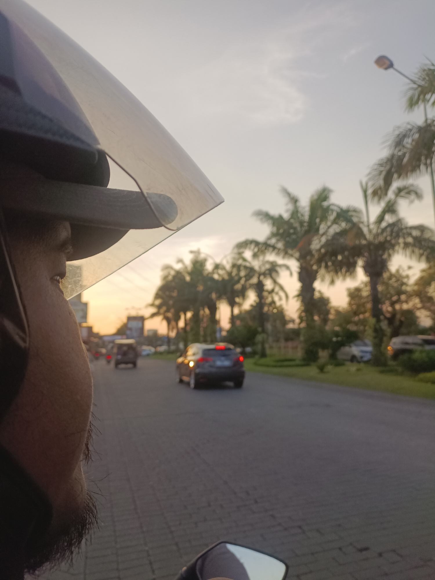 The image depicts a first-person perspective from a motorcycle or scooter, looking forward and slightly to the right. The foreground features the side of a person's face, partially obscured by the visor of a helmet. The person has dark skin, a short beard, and appears to be looking ahead at the road. The helmet is black with a carbon fiber texture on the top, and a clear visor with a slightly reflective surface.
The middle ground shows a paved road with visible brick-like patterns, suggesting it may be a decorative or textured asphalt surface. Several vehicles are on the road. Closest to the viewer on the right side is a dark-colored car, possibly a sedan, with its brake lights illuminated. Further down the road, another dark-colored car is visible in the distance, also with illuminated brake lights. Several other vehicles, appearing as blurry shapes, are spread along the road.
The background is dominated by a warm, hazy sky, indicative of either sunrise or sunset, casting a soft orange glow. Tall palm trees line the right side of the road, with some appearing taller and fuller than others. A street light is visible above the palm trees on the right. The overall environment appears to be an urban or suburban area in Surabaya, Indonesia, suggested by the presence of palm trees, street lights, and the style of vehicles on the road. The lighting and atmosphere suggest a peaceful, possibly evening commute or travel.