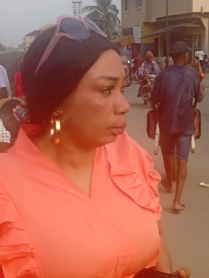 The image captures a candid moment on a busy street, with a woman prominently featured in the foreground, viewed from her right side profile. She has dark, straight hair, and a pair of brown-framed sunglasses are pushed up onto her head. She is wearing large, gold-colored hoop earrings. Her skin tone is warm, and she has visible lines around her eyes and mouth. Her lips are slightly parted, and she appears to be looking intently towards something off-camera to her right. She is dressed in a peach or coral-colored top with a V-neckline and short sleeves that feature ruffled or pleated detailing on the shoulders.
In the background, a bustling street scene unfolds. To the right and behind the woman, a man is seen walking away from the camera, wearing a dark blue t-shirt, dark shorts, and flip-flops. He carries a dark bag or object over his shoulder. Further down the street, several individuals are visible, including at least two people riding motorcycles, one of whom is wearing a patterned top and a light-colored cap or beanie.
Buildings, appearing to be multi-story and somewhat worn, line the street in the distance. A tall, lattice-like structure, possibly a communication tower, is visible in the upper left background. The overall lighting suggests it is daytime, possibly late afternoon or early evening, given the soft, warm light that highlights parts of the scene. The setting appears to be an urban or semi-urban environment.
The brand name "Tom Ford" is faintly visible on the temple arm of the sunglasses worn by the woman.