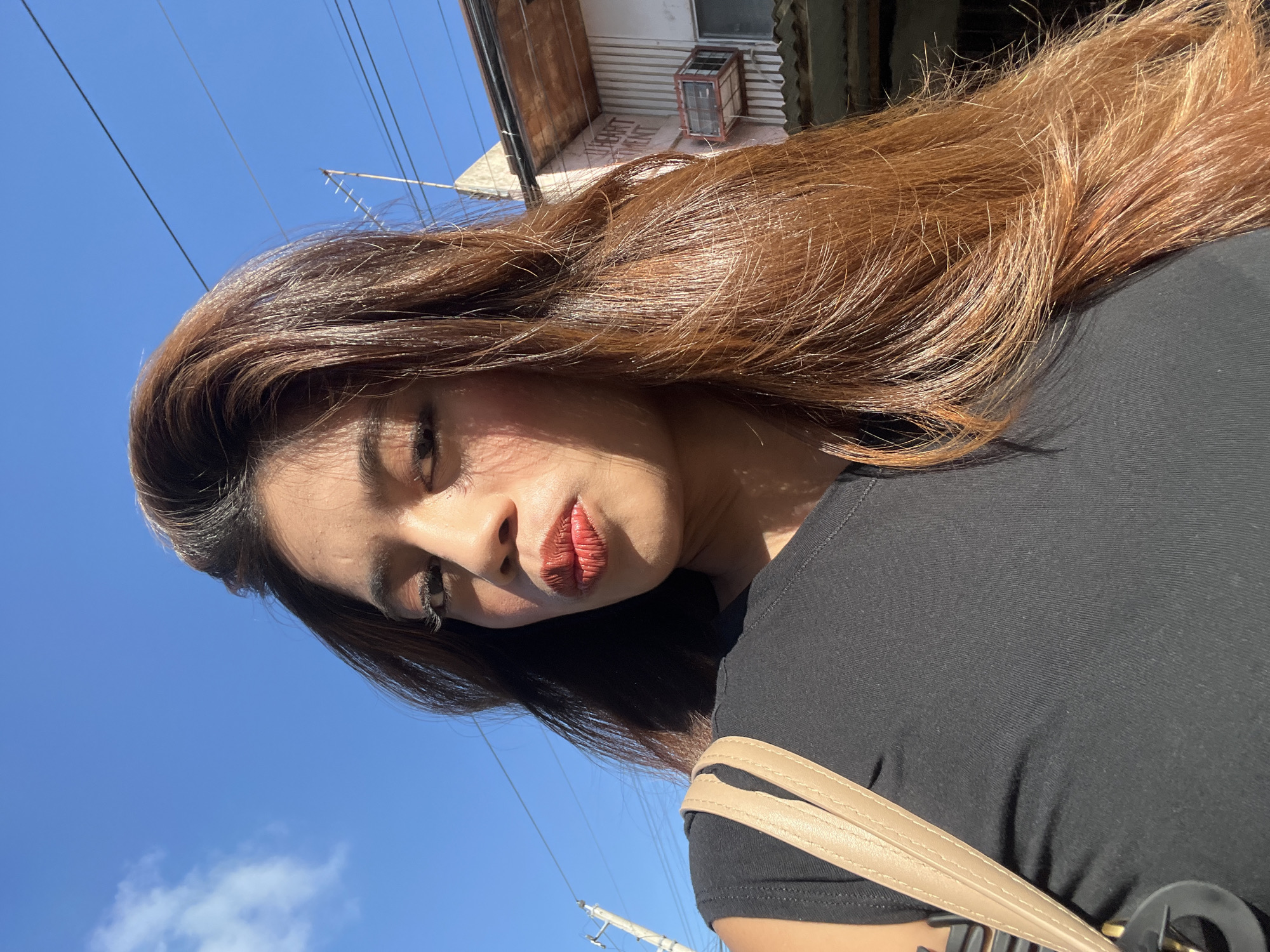 A selfie taken outdoors in Davao City, Philippines. The subject, a woman with long, auburn hair, is the central focus. She is wearing a black top and a tan shoulder bag. Her face is turned towards the camera, with her lips pursed and her eyes looking slightly away. The background reveals a residential area with a building, power lines, and a clear blue sky with a few white clouds. The lighting suggests it is daytime, with shadows cast by the sunlight.