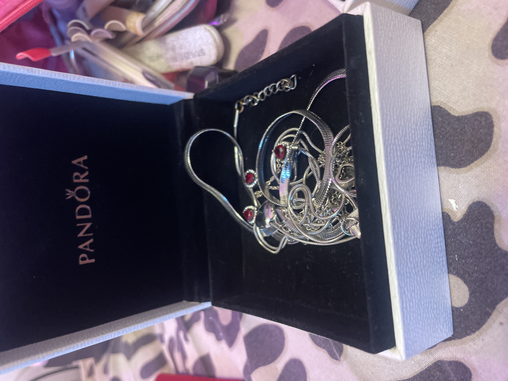 An open Pandora jewelry box, lined with black velvet, contains a tangled silver necklace with two red heart-shaped pendants. The exterior of the box is white with a textured finish. The box sits on a patterned surface. The background is blurred, showing glimpses of other objects, including what appear to be makeup or cosmetic items. The image is taken from a slightly elevated angle. The location in Ladozhskoe Ozero, Russia is not depicted in the image.