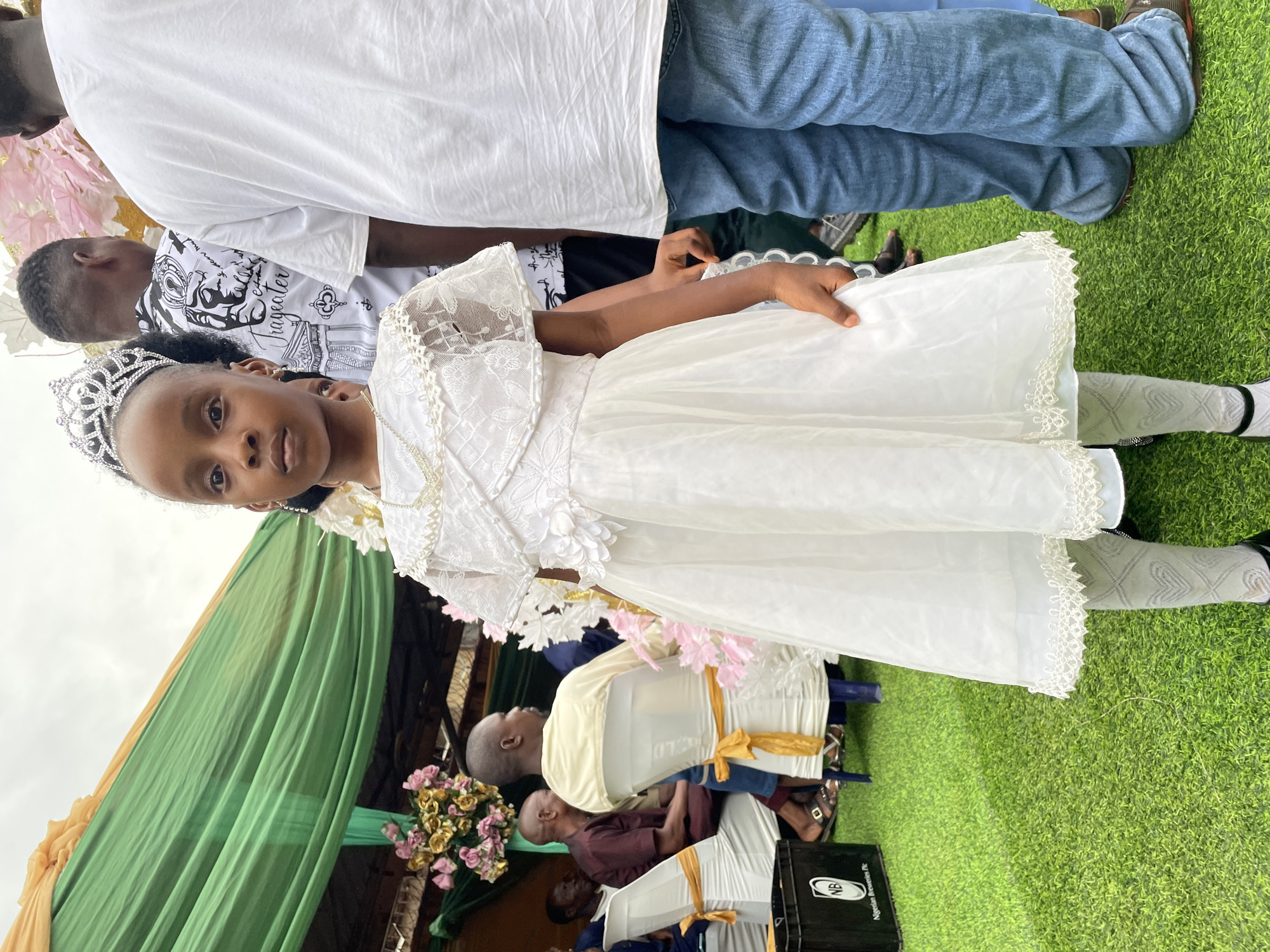 A young African girl, likely between 6 and 10 years old, is the primary subject, wearing a white dress with lace and floral embellishments, a silver crown, and patterned white tights. She stands on green artificial turf.
Behind her, a green and gold draped tent structure is visible, along with seated adults. The presence of decorated chairs, suggests an event like a wedding or religious ceremony.
Other individuals are partially visible, including a person wearing blue jeans and another wearing a white shirt with a patterned black design.
The image was likely taken in Onitsha, Nigeria, judging by the background details.