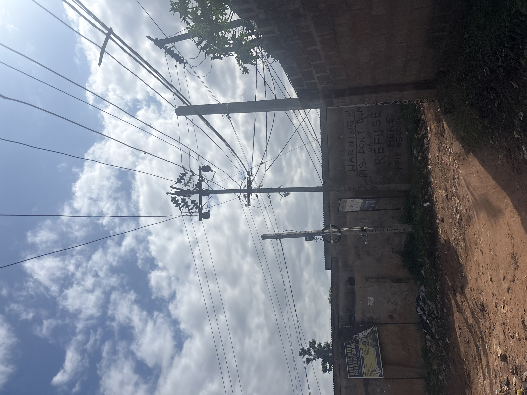 The image depicts a view of a street in Nkpor, Nigeria, with a focus on a wall covered in graffiti. The wall, constructed of brick, serves as a backdrop to a dirt road. Overhead, a complex network of electrical wires crisscrosses the sky, supported by utility poles. On the wall, black spray paint is used to write a warning message: "WARNING !!! PLS. DON'T DUMP UR REFUSE HERE FINE #50000". To the left of the wall, a faded poster is visible, advertising "Kiddies Mind Foundation". The ground is covered in dirt, with scattered debris and vegetation. The sky is partly cloudy, with patches of blue sky visible.
