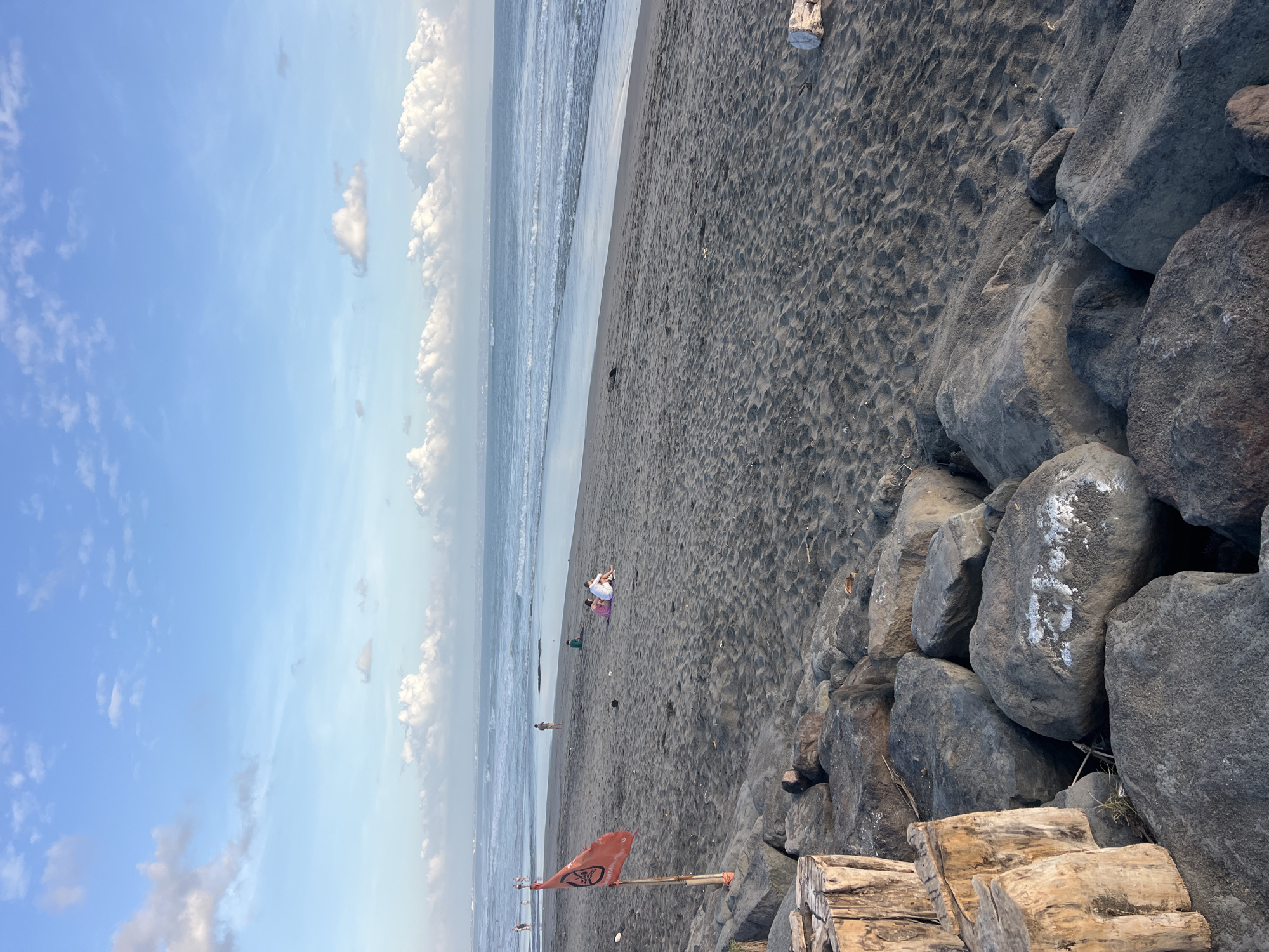 The image depicts a scene at a beach in Mengwi, Indonesia. The foreground features a collection of large, grey rocks, some with white markings, and several weathered wooden posts. Beyond the rocks, the dark sand of the beach stretches towards the ocean. A red flag with a symbol is planted in the sand near the water's edge. In the middle ground, a few individuals are visible on the beach, some sitting and others walking. The ocean is calm with gentle waves breaking on the shore, and the horizon is marked by a hazy outline of land and a clear blue sky with scattered white clouds. The overall lighting suggests it is either early morning or late afternoon.