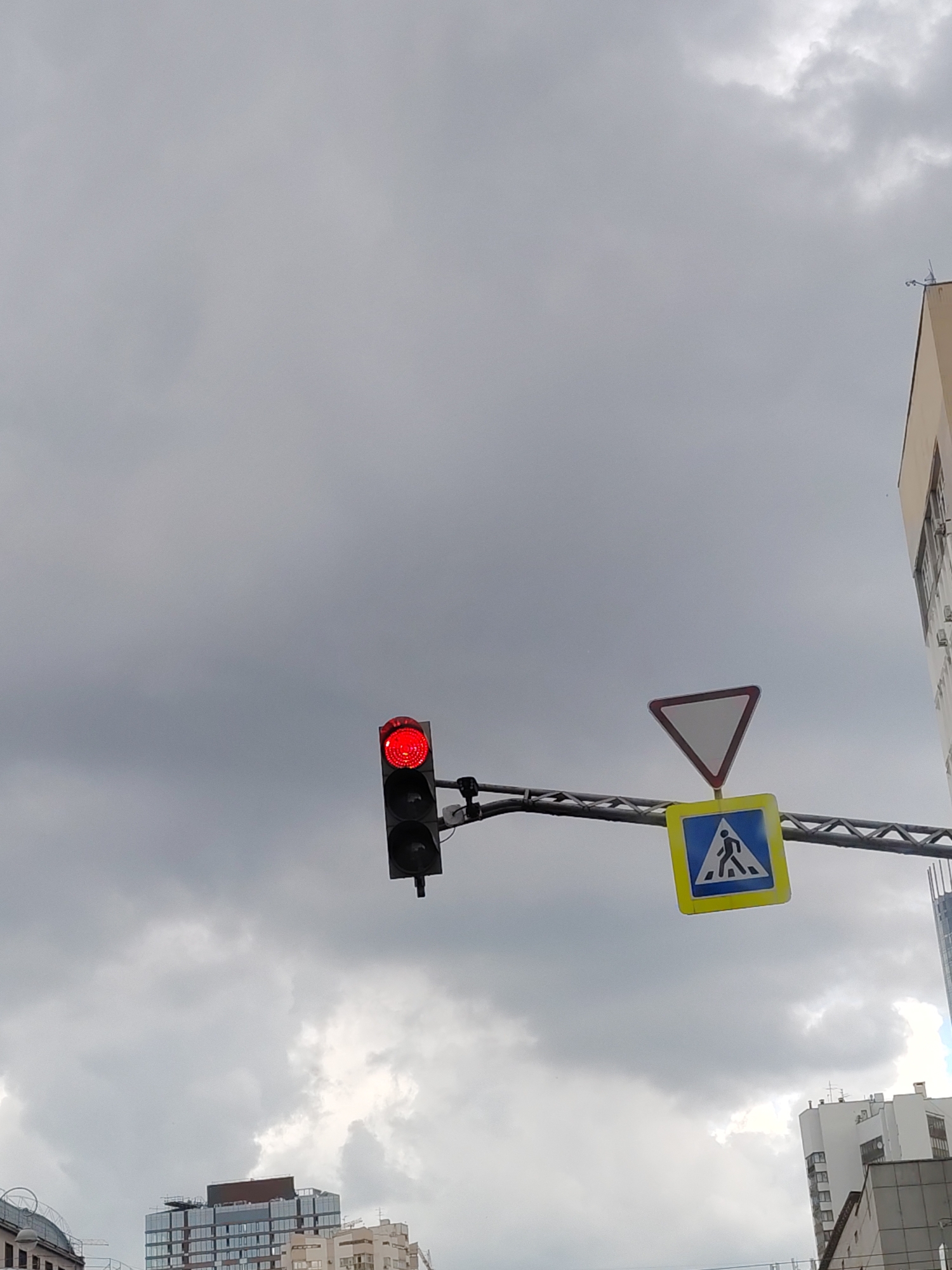 An outdoor, low-angle, vertical shot shows a traffic light with a red signal illuminated, a yield sign, and a pedestrian crossing sign against a cloudy, gray sky. The traffic light is on the left side of the frame. The yield sign, an inverted white triangle with a red border, is positioned to the right and slightly above the pedestrian crossing sign. The pedestrian crossing sign is a yellow square with a blue inner square, displaying a white silhouette of a person walking. The signs are mounted on a horizontal metal beam. In the background, the tops of several multi-story buildings are visible, including a modern glass-fronted building on the right, with lighter colored buildings below and to the left. The overall scene suggests an urban environment with overcast weather. This scene is located in Yekaterinburg, Russia.