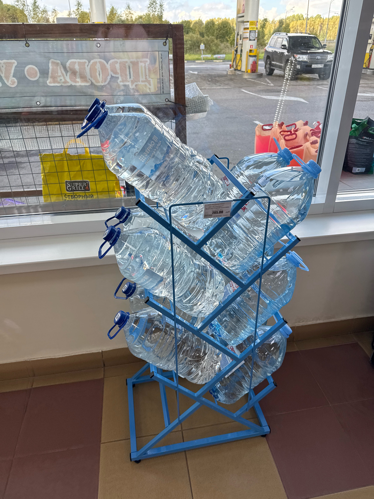 The image shows a display of large plastic water bottles arranged on a blue metal rack. The bottles are clear and appear to be filled with water. Some of the bottles have blue caps and attached handles. A price tag is visible on one of the bottles, indicating "285.00 руб." The display is positioned indoors, near a large window, through which a gas station and a black SUV are visible. The exterior scene suggests an outdoor location, possibly a service station or convenience store, given the presence of gas pumps. The floor inside appears to be tiled. The overall impression is a retail environment, likely a store within a gas station, selling bottled water.