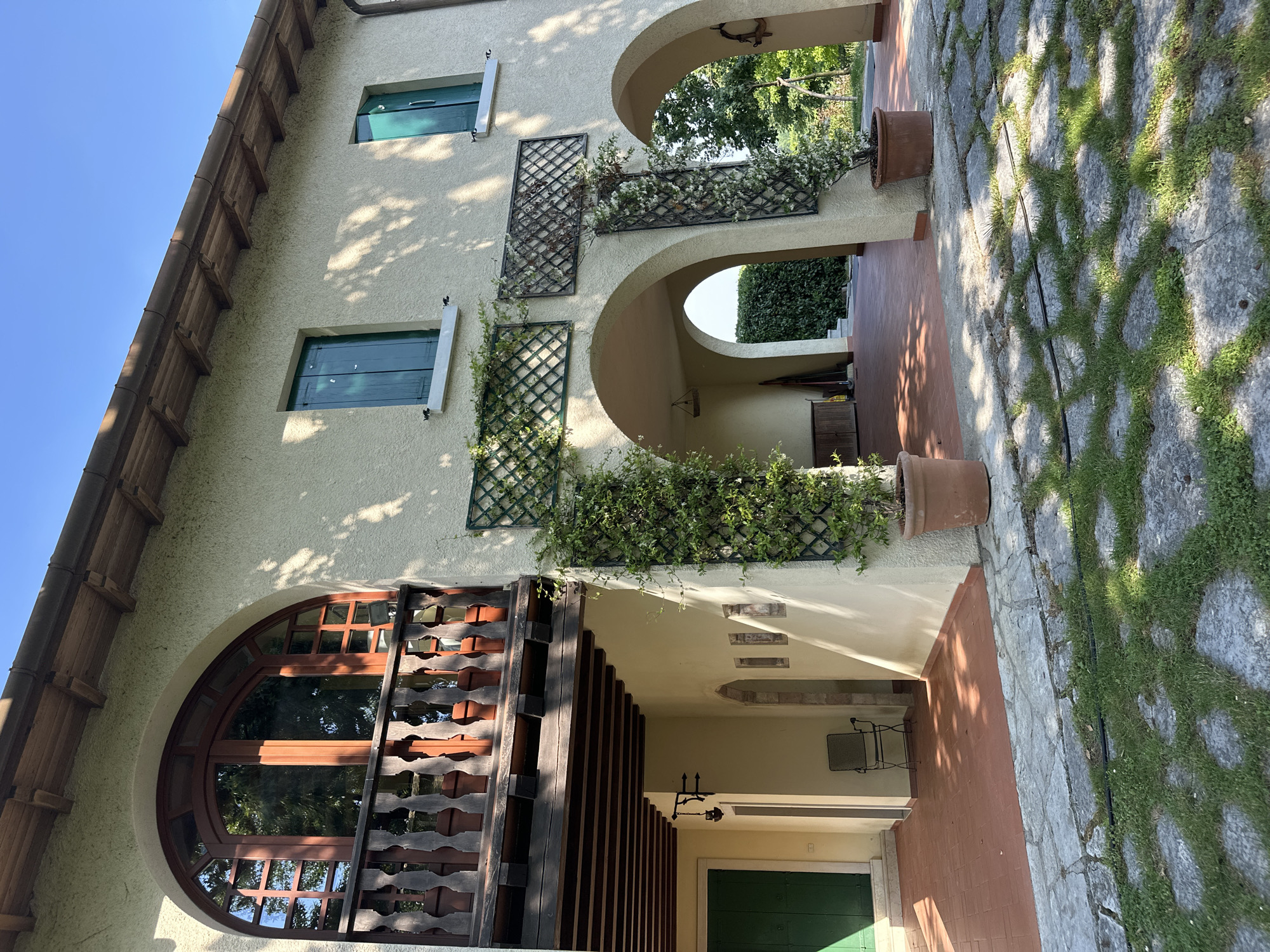 This media file shows a building with a light-colored stucco exterior and wooden architectural details. The building features several arched openings, some of which are covered with trellises supporting climbing plants. There are at least two windows with green shutters. A large, arched window with wooden framing is visible on the left side of the image. A walkway paved with stone and grass leads to the building, with terracotta pots containing plants placed along the path. The scene suggests a sunny day with dappled shadows cast by trees. The architecture and landscaping hint at a Mediterranean or Italian setting, consistent with the provided location context of Conegliano, Italy. No people or specific activities are depicted. The image appears to be taken from a low angle, looking up towards the building. There is no visible text in the image.