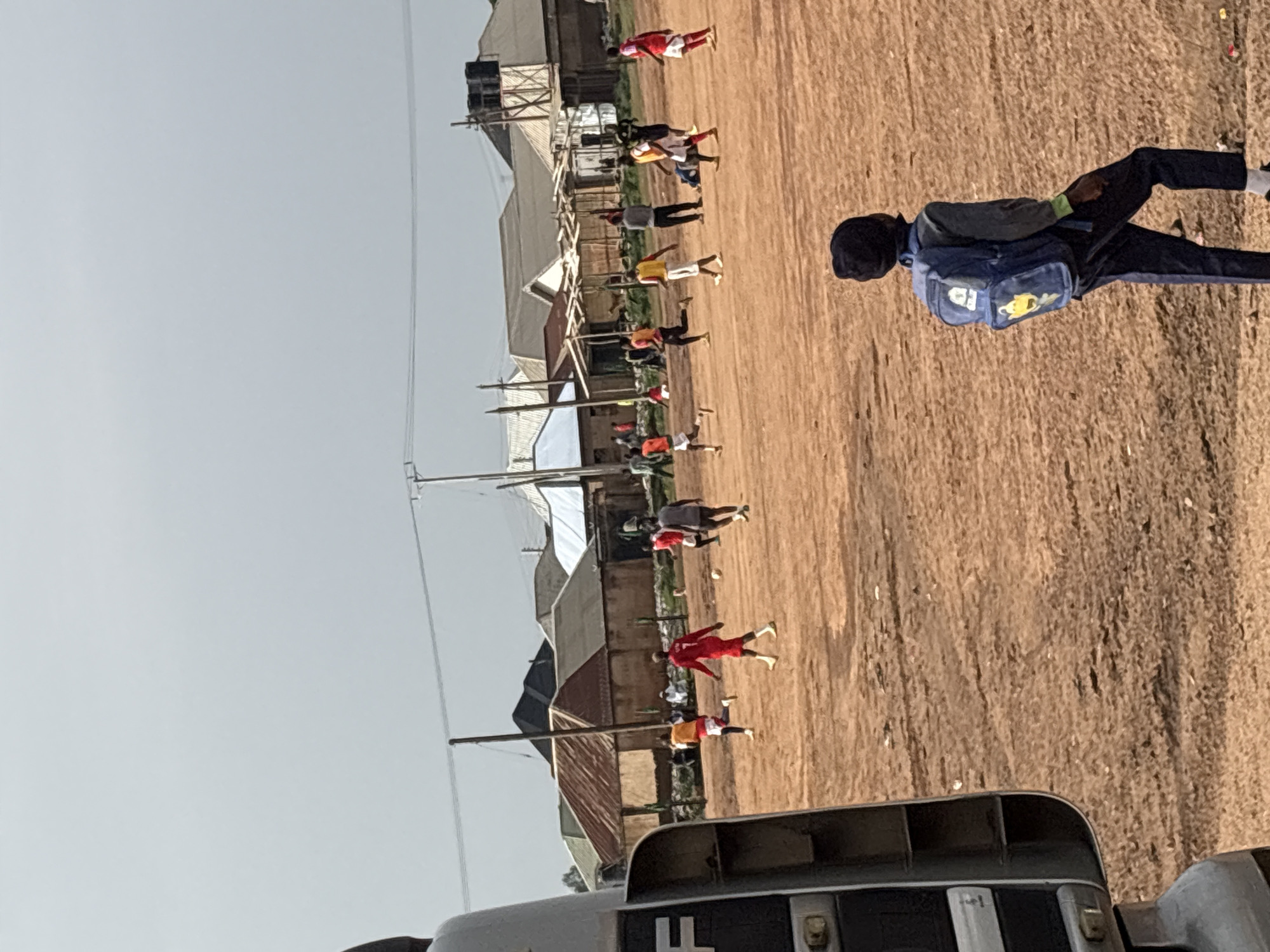The image shows an outdoor scene in what appears to be Bukuru, Nigeria. A line of people is seen in the distance, some dressed in sportswear, possibly exercising or training. Buildings and utility poles are visible in the background, suggesting an urban or peri-urban environment. A child carrying a blue backpack walks on the right side of the image. A vehicle is partly visible at the bottom. The ground is brown and appears to be dirt or sandy soil. The sky is overcast.