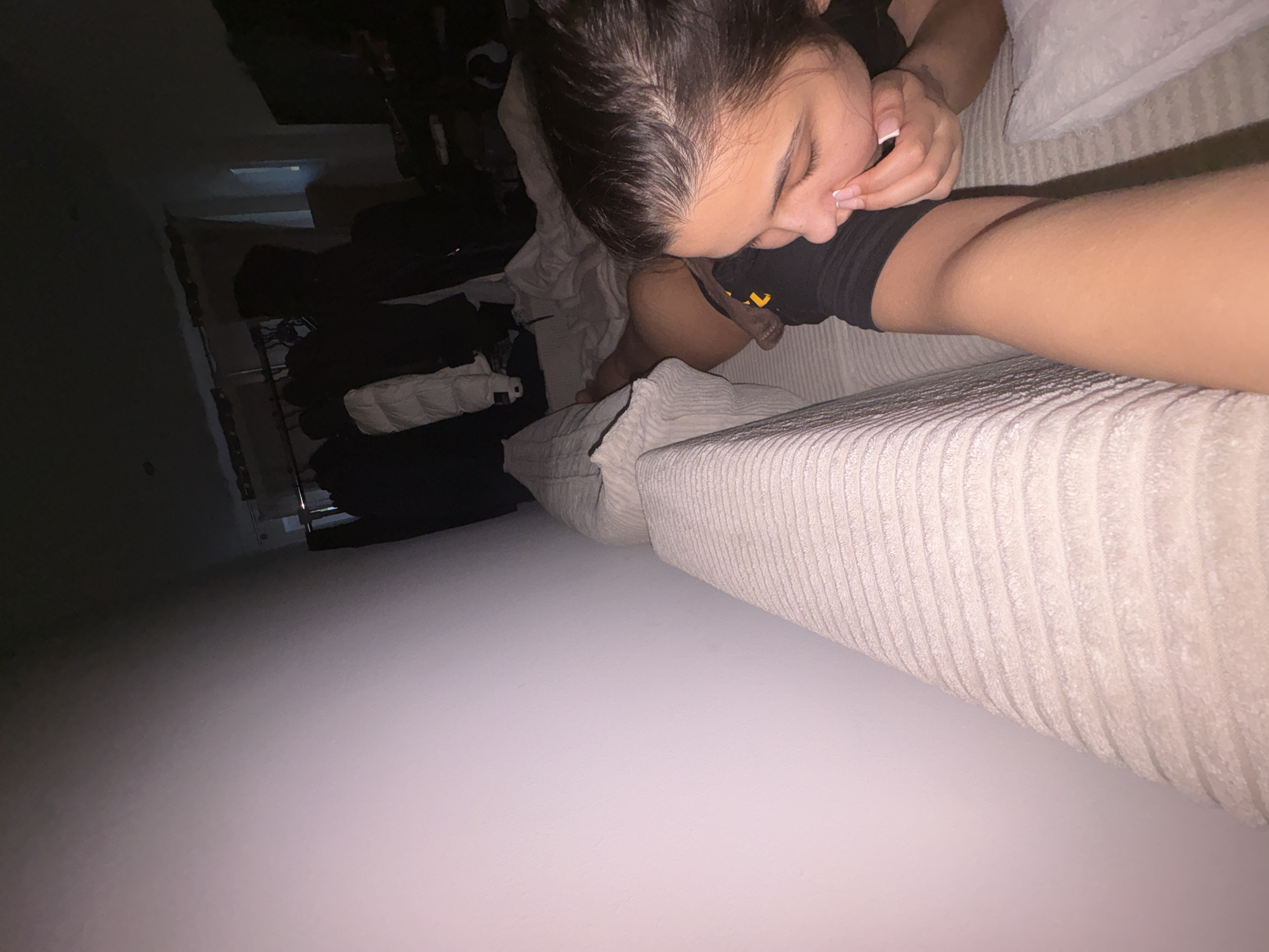 A close-up, eye-level shot captures a young person lying on a textured couch in what appears to be a bedroom. The person, with dark hair and wearing a black shirt, is looking down and touching their face with their fingers, particularly their nose. Their gaze is directed towards their own leg, which is extended in front of them and partially visible. The surrounding environment is dimly lit, with a wall on the left side of the frame. In the background, clothing items are hanging on a rack, suggesting a closet area. The overall impression is that of a private, intimate moment. This scene is taking place within a private residence, an apartment or house, in Mönchengladbach, Germany.