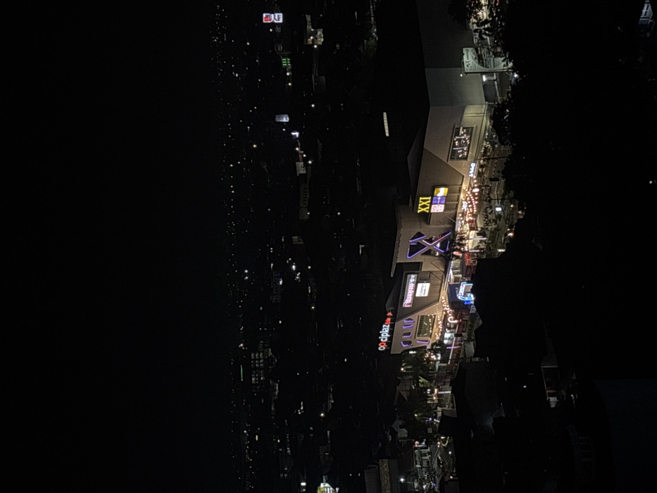 An aerial nighttime view depicts a brightly illuminated commercial complex, identified by prominent neon signs for "ciplaz," "Transmart," and "XXI" cinema, situated within the city of Sidoarjo, Indonesia. The complex features modern, angular architecture with multiple levels. An outdoor area directly in front of the main entrance is lit by numerous string lights, beneath which a gathering of individuals is observed. Vehicle lights are visible on the street immediately adjacent to the complex. The surrounding environment is an urban landscape, extending into the dark background as a sprawling network of scattered residential and commercial lights beneath a clear, starless sky. No specific interactions are discernable, but the presence of many people indicates public activity.