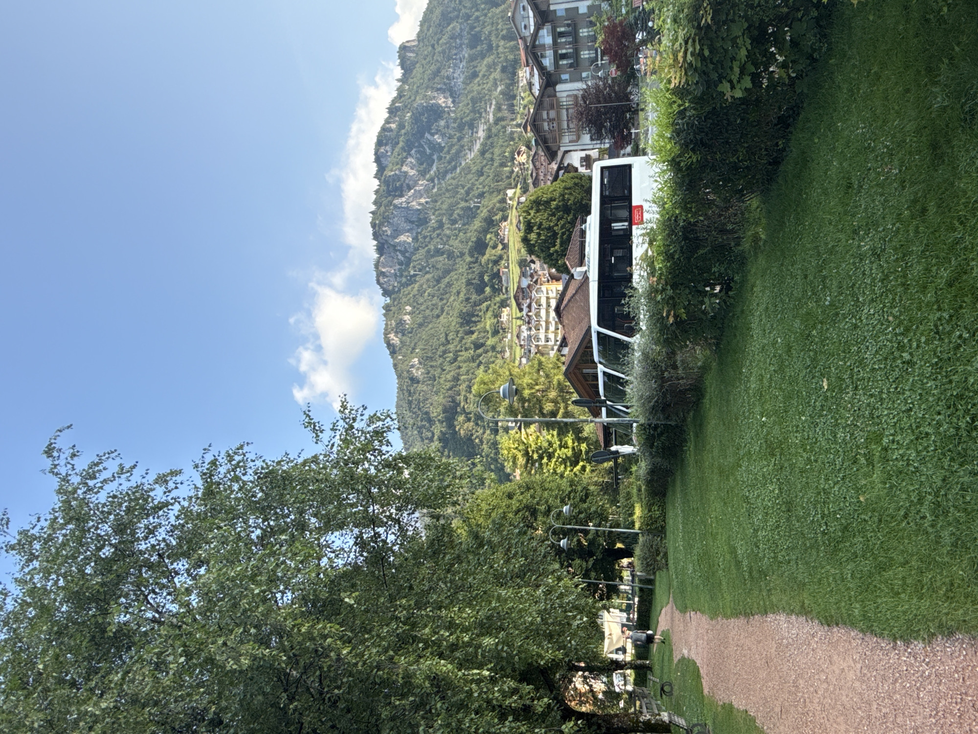 A daytime, eye-level, full shot captures a pathway and grassy area leading towards a mountain village in Molveno, Italy. In the foreground, a light brown gravel path curves to the left, bordered by lush green grass. A lone figure, dressed in a dark t-shirt and shorts, walks away from the camera on the path. Beyond the grassy area, a white bus with tinted windows and a red logo is parked. Behind the bus, a dense hedge separates the foreground from a street lined with buildings and trees. In the background, the village of Molveno is nestled at the base of a rugged, tree-covered mountain, with homes and buildings scattered across the slope under a bright blue sky with scattered white clouds. The scene conveys a serene and picturesque atmosphere of a European mountain town.