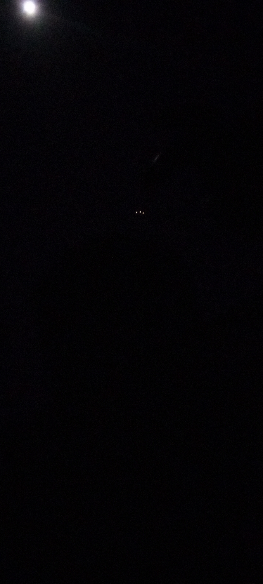 The image presents a predominantly dark, nocturnal scene, featuring minimal light sources against a black background. In the upper-left quadrant, a bright, diffused white light is visible, likely representing the moon due to its intensity and halo effect, though it appears overexposed. Towards the center-right, three small, distinct, yellow-orange lights are horizontally aligned, suggesting artificial illumination from a distant source such as a vehicle or structure. Faint, elongated streaks of light are subtly visible in other dark areas, possibly indicating reflections or very distant light trails. No other discernible objects, people, or activities are present. The pervasive darkness clearly indicates a nighttime setting. There are no specific visual cues within the frame to confirm the location as Ilogbo Eremi, Nigeria, nor is any text visible.