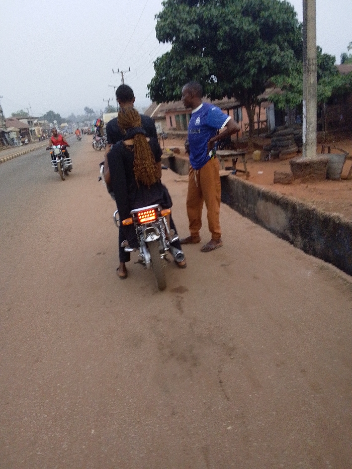 Two people on a parked motorcycle face away from the camera, stopped on the side of a road. A man wearing a blue shirt and khaki pants stands next to them, his hands on his hips. Other motorcyclists are on the road ahead. Buildings and trees line the roadside. The sky is overcast. The scene seems to be a rural or suburban setting.