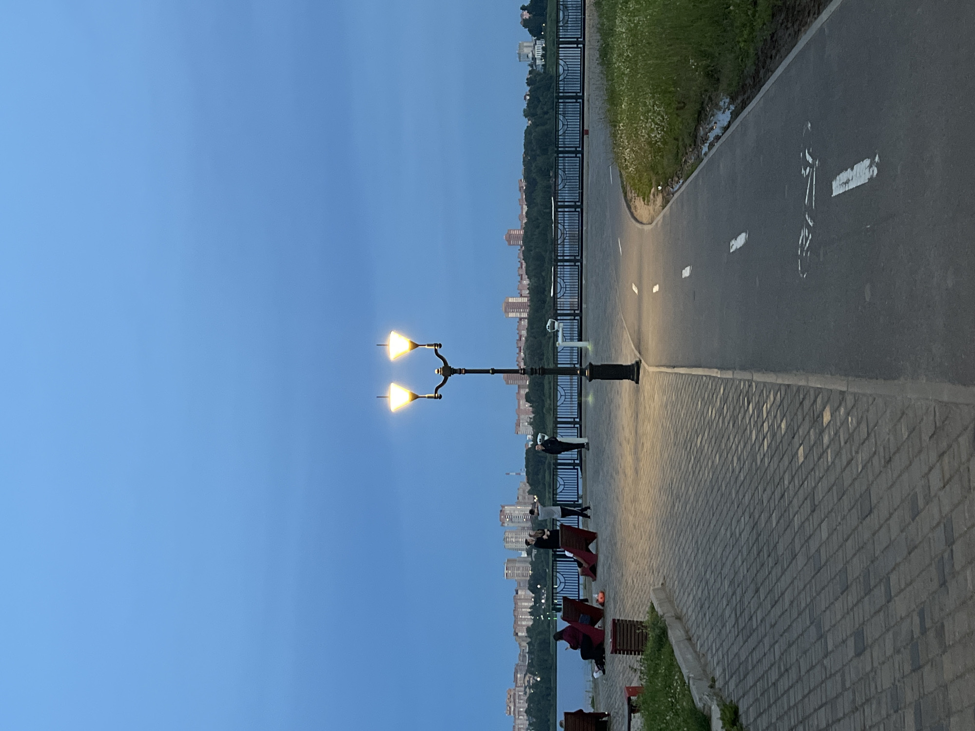 The image depicts a waterfront promenade in Cherepovets, Russia, during twilight. A prominent double-headed lamppost illuminates the paved walkway and a separate asphalt bicycle path. In the background, the calm waters of the Sheksna River are visible, bordered by a black metal fence and a tree line. Across the river, a city skyline with apartment buildings and some scattered trees is silhouetted against the deepening blue sky. On the promenade, several individuals are present. Two people are seated on red benches, facing the river. Further along, a group of four people are gathered near the fence, with two of them appearing to be in conversation or interaction. Another person stands alone on the promenade to the right of this group. The bicycle path is marked with white lines and a painted bicycle symbol. The foreground shows the textured paving stones of the walkway leading towards the lamppost. Green grass and some wildflowers border the bicycle path on the right. The overall scene suggests a serene evening at a public recreational area.