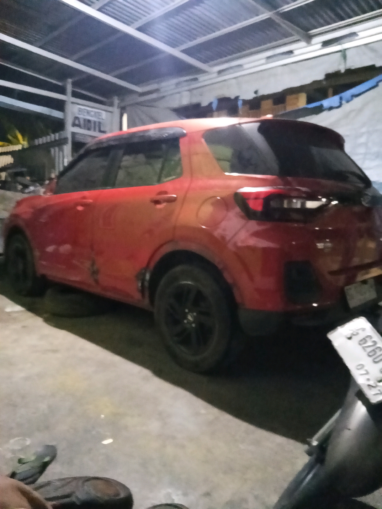 A red SUV is parked indoors, possibly in a garage or workshop. The car is red and has black rims on its wheels. There is a sign in the background that reads "BENGKEL AMIL". A license plate with the number "6260" and "07-27" is visible in the foreground, partially obscuring a motorcycle. The environment appears to be a concrete floor with some debris scattered around. The lighting is dim, suggesting it is either nighttime or indoors with limited light.