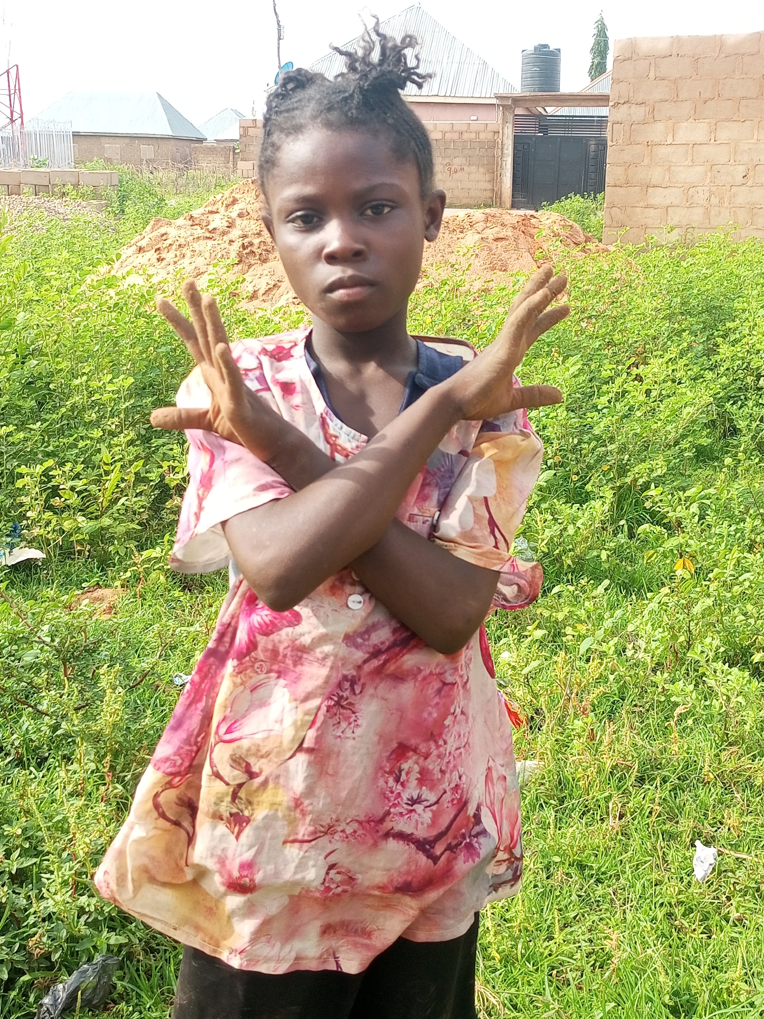 A young girl stands outdoors in a grassy area in Tempure, Nigeria. She is wearing a light-colored dress with a floral pattern in shades of pink and yellow. Her arms are crossed in front of her chest, with her hands held up, fingers spread. The background shows construction materials, including a pile of sand and brick walls, suggesting an ongoing building project. A water tank is visible on top of a structure in the distance. The ground is covered in green vegetation, and there are some scattered pieces of litter. The lighting indicates it is daytime, with clear skies.
