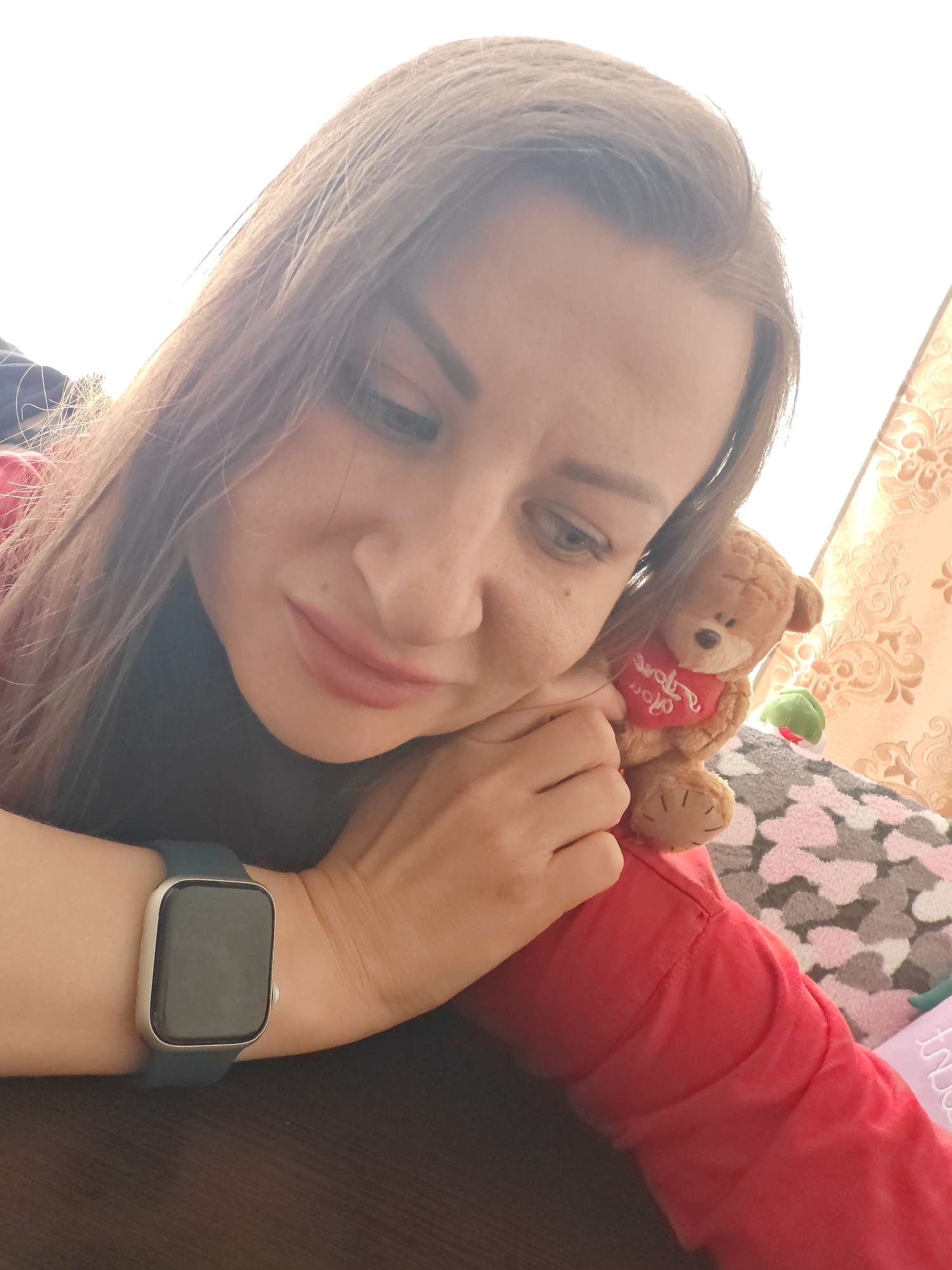 This is a close-up, over-the-shoulder shot of a woman holding a small teddy bear. The woman has long, dark brown hair that is slightly blurred due to the shallow depth of field. She is wearing a red garment, and on her left wrist, she has a smartwatch with a dark band and a blank screen. The teddy bear is light brown and has a red heart-shaped bib around its neck with some white text on it. The woman's hand is gently holding the bear, and she is looking down at it with a soft expression.
The background is out of focus but appears to be an indoor setting. There is a patterned fabric, possibly a blanket or upholstery, with a pink and grey floral design. To the right of the woman, a portion of a wall with a textured, possibly wallpapered, pattern is visible. The lighting suggests it is daytime, with bright light coming from the upper left, casting a soft glow on the woman's hair and face. The overall mood of the image is tender and intimate, conveyed by the woman's gentle interaction with the teddy bear and her soft gaze. The text on the teddy bear's bib is too blurry to read.