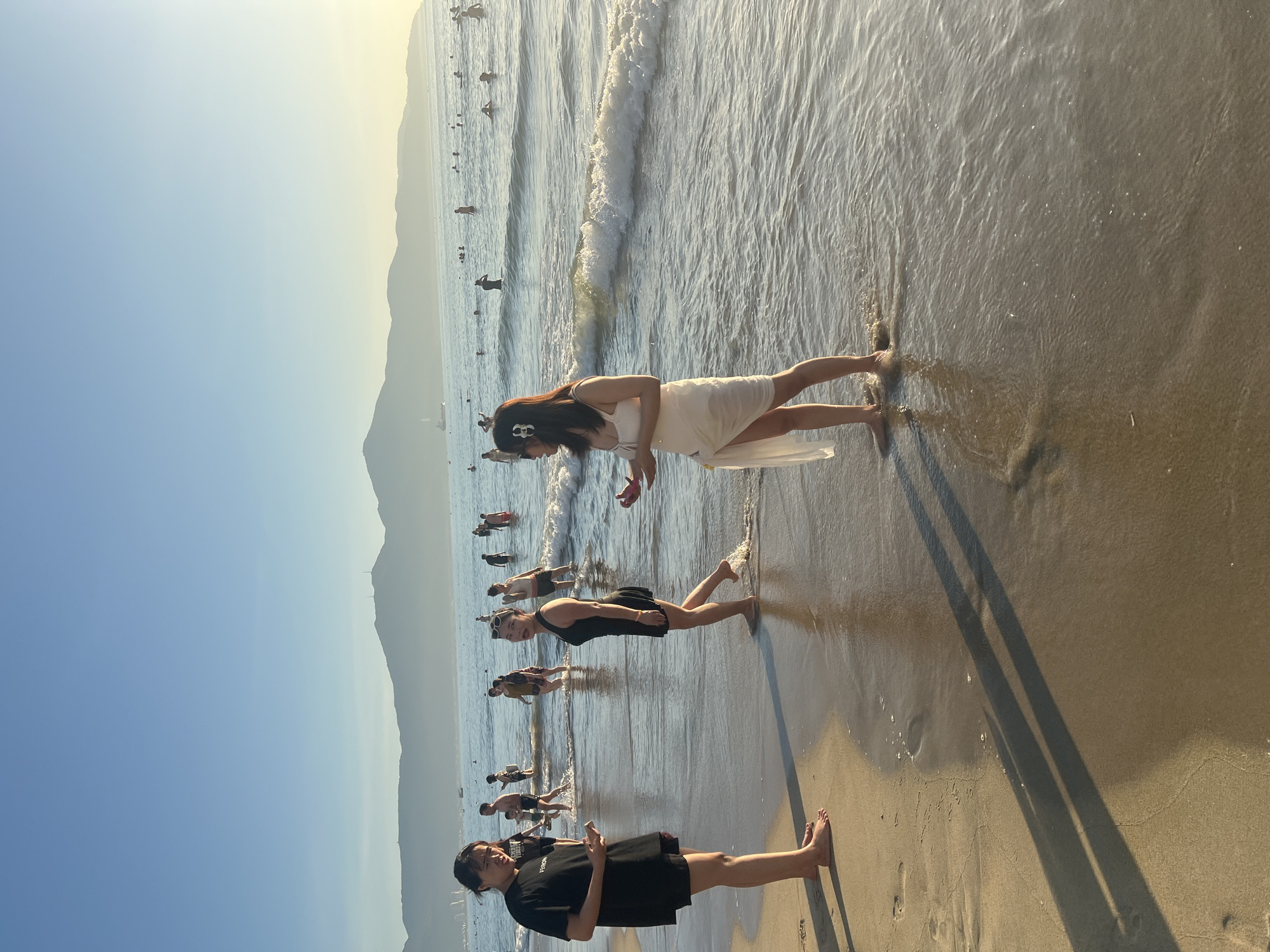 The image depicts a beach scene in Da Nang, Vietnam, with a clear sky and distant mountains. Several people are visible, some wading in the shallow ocean water and others on the sandy shore. In the foreground, two women are standing on the wet sand. The woman on the left, wearing a black t-shirt and shorts, is holding a mobile phone and looking at the woman to her right. The woman on the right, dressed in a white cover-up over a black swimsuit, is looking towards the ocean and has her hand near her mouth. Further out in the water, a crowd of people are swimming and standing. The sun appears to be setting or rising, casting long shadows on the sand and creating a warm, golden light across the scene. Waves are gently breaking on the shore, indicating a calm sea.
