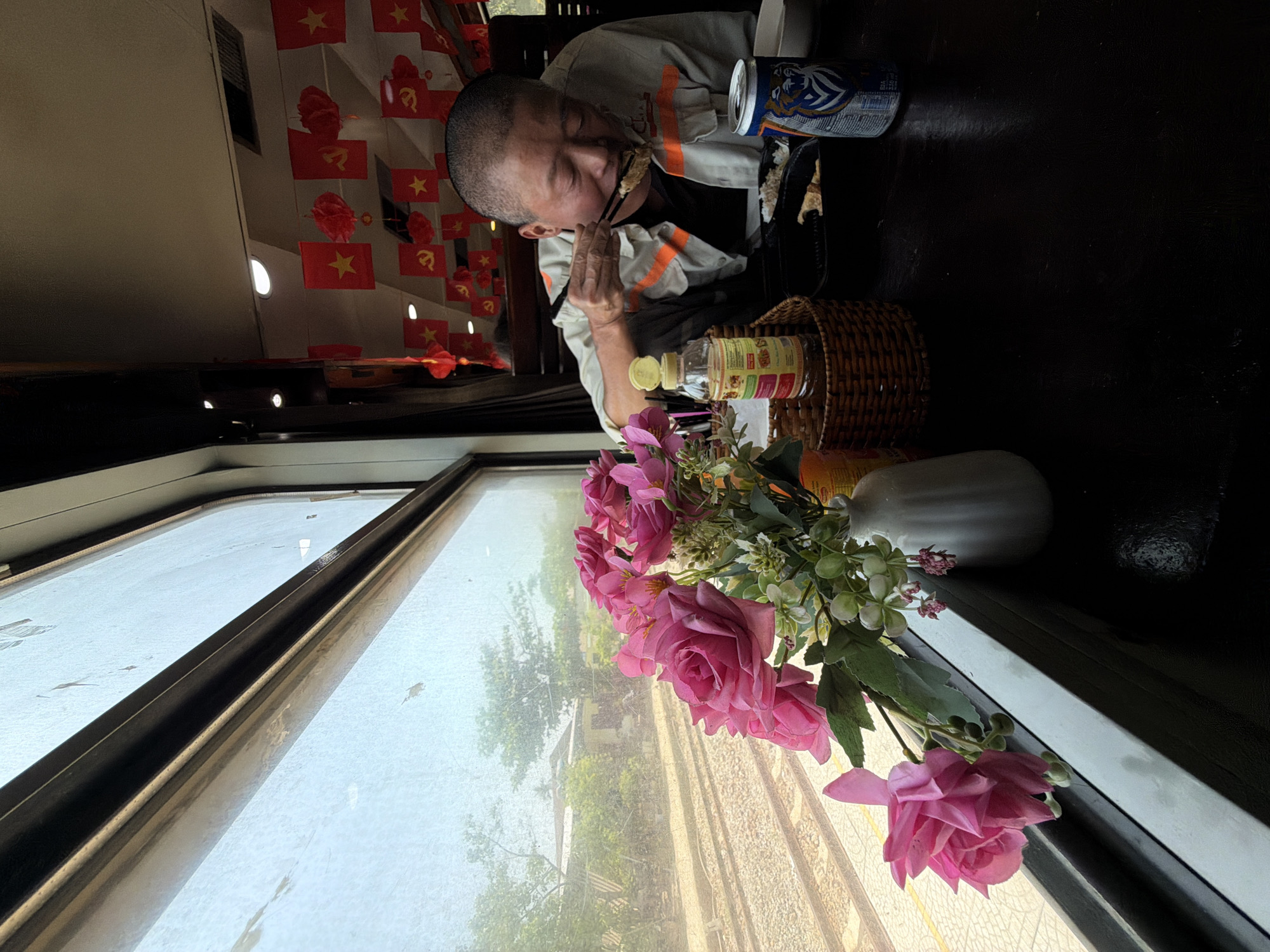 An interior scene depicts a man of Asian descent seated at a dark table in what appears to be a train car, as evidenced by the window showing moving tracks. He is using chopsticks to eat. A vase of pink flowers sits by the window, alongside a bottle of liquid in a woven basket. A can of beer rests on the table near the man. Red flags with yellow stars are visible, suggesting the location is within Vietnam. Based on this information, it's likely that the location is in the city of Huế, Vietnam.