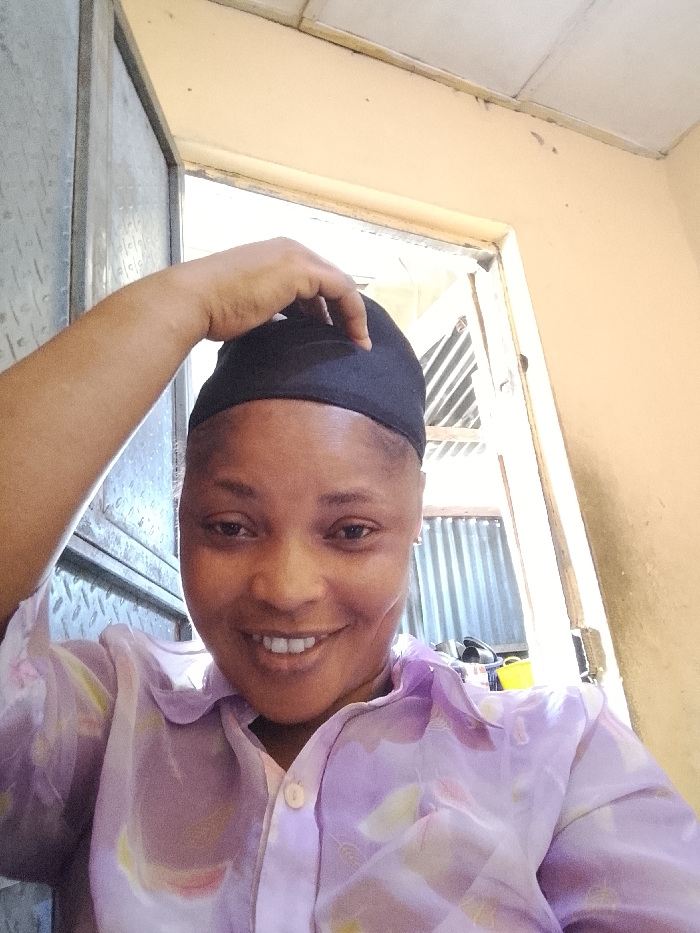 A smiling woman wearing a light purple, floral-patterned button-down shirt and a black head covering poses indoors. She is touching the top of her head covering with her right hand. Behind her is a doorway, showing a metal-corrugated structure. The background also includes an off-white wall and a partially visible window.
