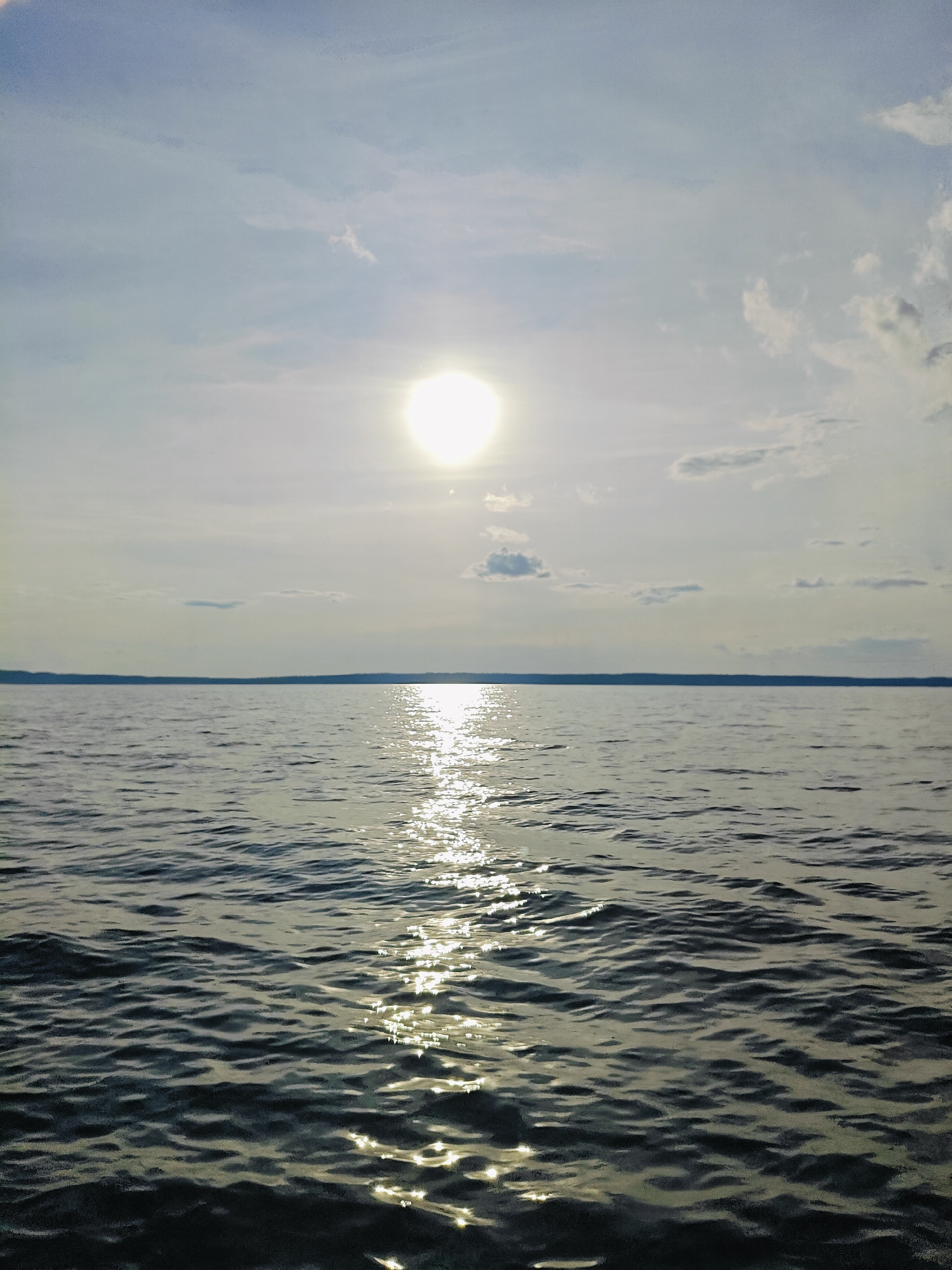 A wide shot captures a calm body of water under a hazy sky. The sun is high and bright, its rays reflecting off the water's surface in a shimmering path. A distant, dark shoreline stretches horizontally across the frame, suggesting the water is a large lake or river. The sky is a pale blue, dotted with scattered clouds, some wispy and translucent, others more defined. The overall impression is one of serene, open space, possibly a view from a boat or the shore of the Kama River near the city of Chaykovsky, Russia, known for its expansive water views. There are no people or objects in the immediate foreground.