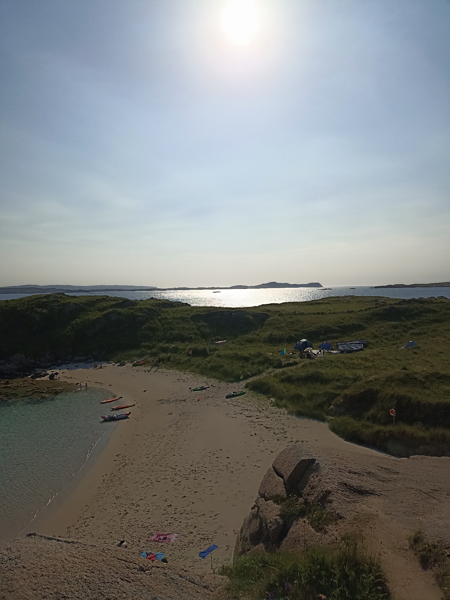 The image depicts a serene coastal landscape on a bright, sunny day. In the foreground, a sandy beach slopes down to clear, shallow water. Several kayaks are resting on the sand and in the water, suggesting recreational activity. Scattered towels add a touch of color to the beach.
Further inland, rolling green hills dotted with patches of grass and rocks dominate the scene. A small campsite is visible on one of the hills, featuring several tents and a small boat. In the distance, the calm, sun-drenched ocean stretches out, meeting the horizon where islands can be seen. The sun is high in the sky, casting a bright glare on the water.
The overall impression is one of a peaceful, natural environment, likely a secluded bay or cove, ideal for outdoor activities like camping and kayaking. The scene captures the beauty of a coastal summer day. The location is specified as Carnboy, Ireland.