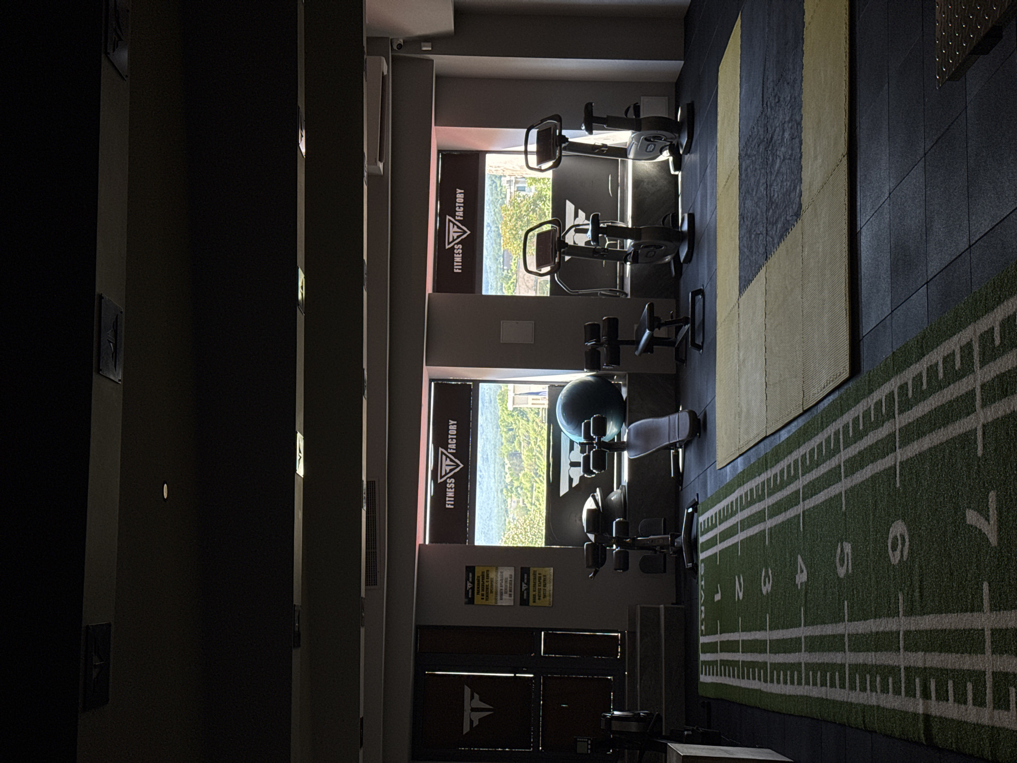 The image depicts the interior of a fitness center located in Tsarevo, Bulgaria. A row of exercise equipment, including stationary bikes and weight machines, is positioned along a wall with large windows displaying an outdoor view of what appears to be a coastal landscape. Banners with the logo "Fitness Factory" are visible in the windows. The floor is covered with black and yellow exercise mats, and a green artificial turf with white yard lines and numbered markings is present, resembling a football field. The interior is brightly lit from the daylight entering through the windows. The image is taken from a slightly elevated and angled perspective, and there are no people visible in the scene.