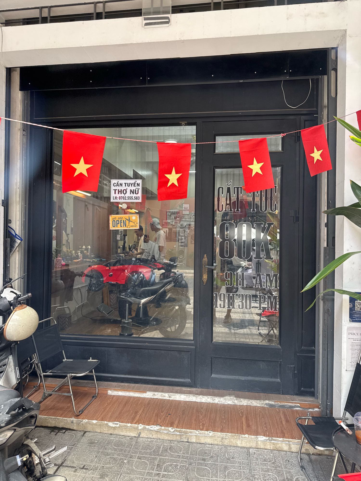 The image shows the exterior of a barbershop in Nha Trang, Vietnam, as indicated by the Vietnamese text and the presence of Vietnamese flags. The shop has a large glass storefront with a black frame, allowing a view of the interior. Inside, a barber chair and styling equipment are visible, suggesting a functional barbershop. Two men are present inside, one appearing to be a customer and the other a barber. Outside, in front of the shop, there are several motorcycles parked, which is common in Vietnamese cities. A red toy car is also visible inside, potentially for children. Vietnamese flags are strung across the front of the shop. Signage on the door indicates "CẮT TÓC 80K," meaning "Haircut 80K," and specifies operating hours from 9 AM to 7:30 PM. Another sign says "CẦN TUYỂN THỢ NỮ" which translates to "Female Barber Wanted." The overall scene depicts a typical street-level business in Vietnam.