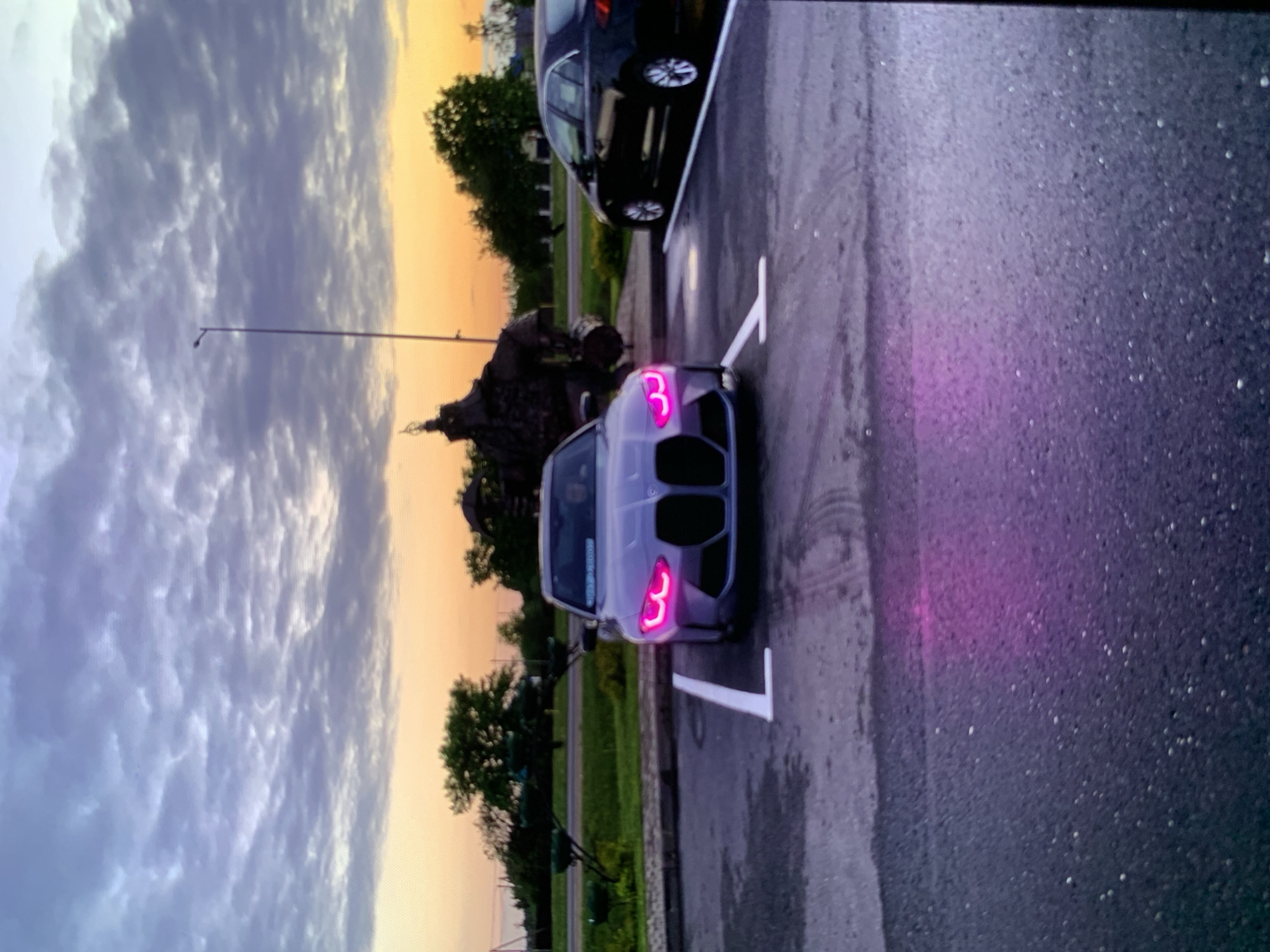 An eye-level, medium shot captures a gray BMW M3 sedan parked in a parking lot under a cloudy sky with the sun setting in the background. The car's headlights glow with a vibrant pink hue, and a pinkish glow also reflects off the wet asphalt in front of the car. The car is parked in a marked spot, indicated by white lines on the ground. To the right of the BMW, another car, a dark-colored sedan, is parked in an adjacent spot. In the background, trees and a sculpture are visible. The overall scene is one of a car parked in an outdoor lot during twilight.