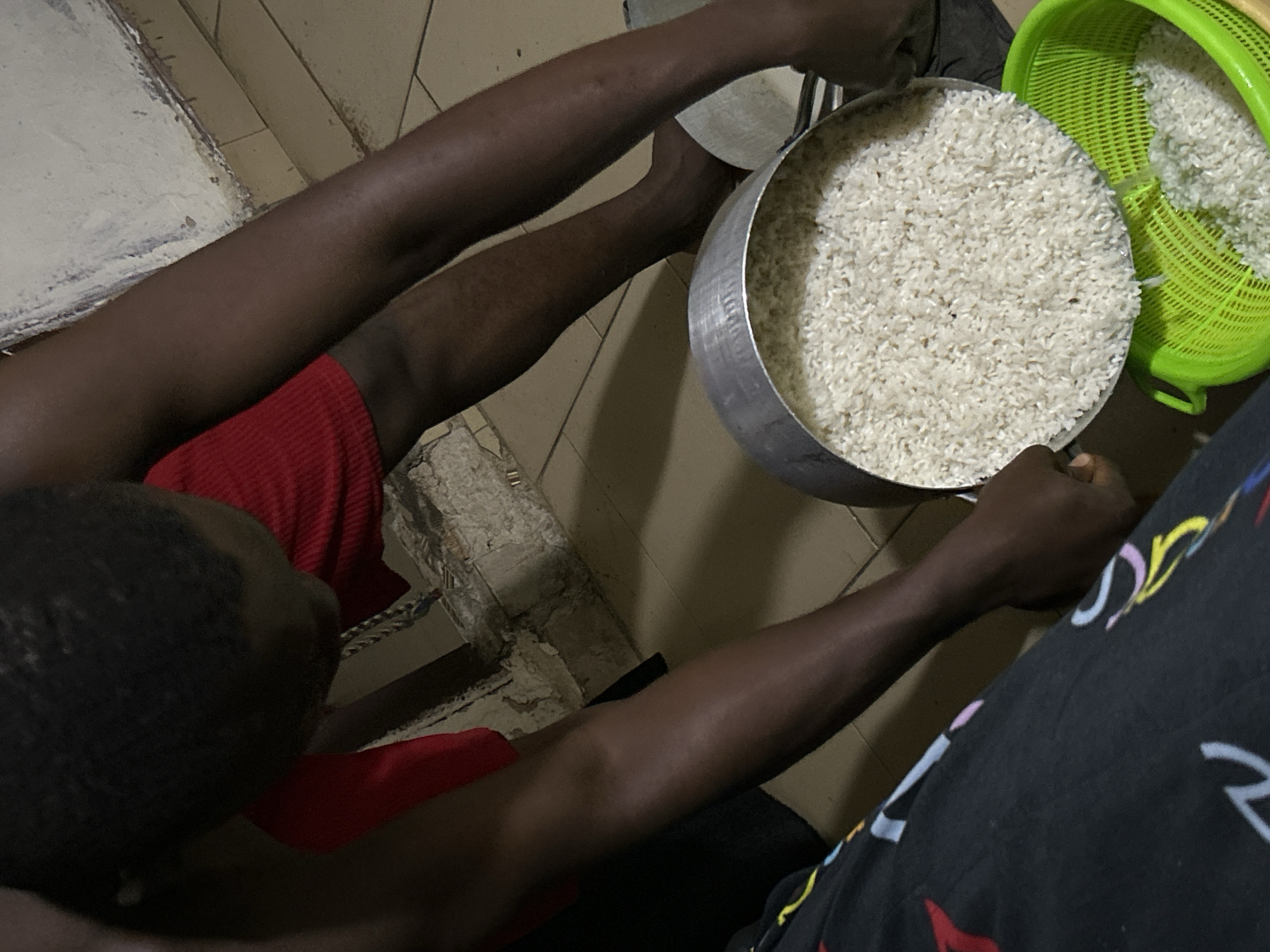 This media file shows a close-up, overhead view of two people and a large amount of uncooked white rice. The primary subject is a person wearing a red garment, whose dark-skinned legs and arms are visible. They are holding a metal pot filled with rice. To the right of this pot, a bright green plastic colander also contains rice. The scene appears to be indoors, possibly a kitchen or dining area, with tiled flooring visible. The lighting suggests it might be evening or a dimly lit indoor space. The overall mood is neutral, focusing on the task of handling the rice. There is no visible text in the image. The context provided places this scene in Ihiagwa, Nigeria.