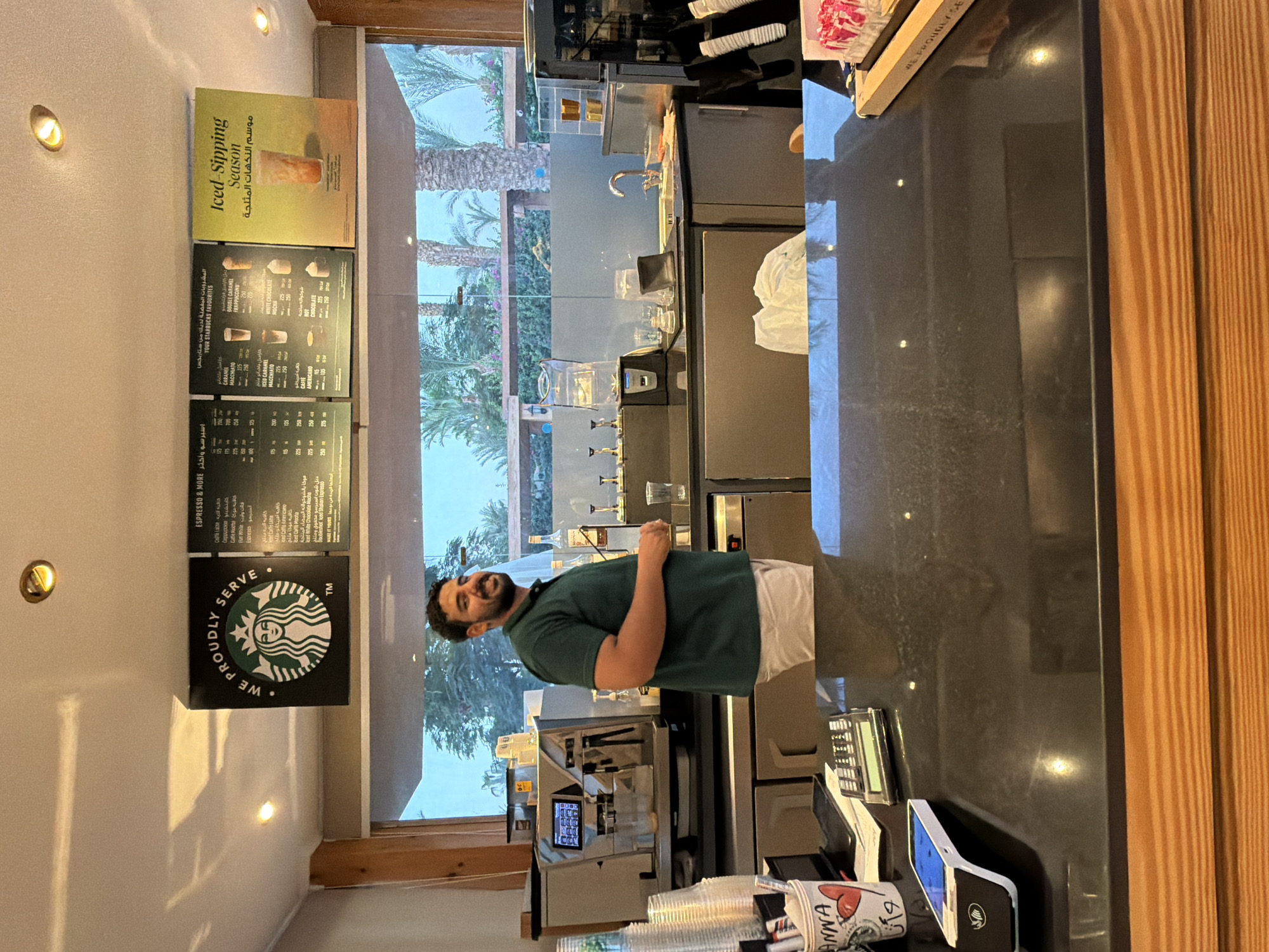 The image depicts the interior of a Starbucks coffee shop, likely in Sharm El Sheikh, Egypt, given the presence of Arabic text on the menu boards and promotional materials. A male barista, with dark hair and a beard, wearing a dark green polo shirt and white pants, stands behind the counter, facing the camera and smiling. He is holding what appears to be a receipt or small slip of paper in his right hand.
The counter is made of dark, reflective material, with a cash register, a credit card reader, and a cup with "ANNA" written on it visible on the left side. Behind the barista, a commercial espresso machine is positioned. The background features a large window overlooking an outdoor area with palm trees, suggesting a warm climate.
Above the counter, a prominent Starbucks logo is displayed, along with menu boards showing drink options in both English and Arabic. One promotional sign features the text "Iced-Sipping Season" in English and its Arabic translation. Various coffee-related equipment, cups, and bottles of syrups are visible on shelves and the counter in the background. The overall environment appears clean and functional, typical of a coffee shop setting.