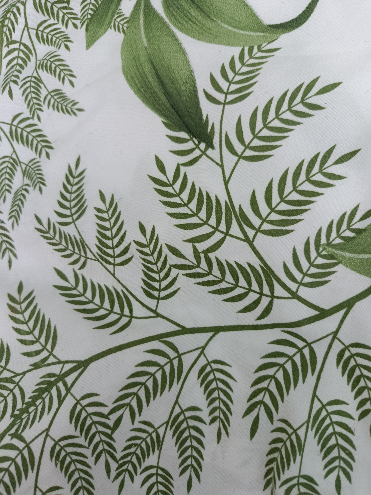 The image displays a close-up of a white fabric with a repeating pattern of green leaves and branches. The leaves are depicted in various sizes, with some larger, more solid green leaves towards the top right, and smaller, more delicate fern-like leaves forming branches. The pattern has a slightly textured appearance, suggesting it might be printed on cotton or a similar material. The lighting appears even, and there are no shadows indicating a specific light source. The scene is devoid of any people, animals, or architectural elements, making it impossible to ascertain its location within Tashkent, Uzbekistan, or any other city. The image solely focuses on the textile design.
