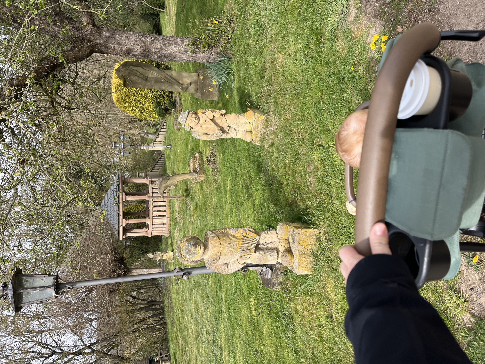 The image shows an outdoor scene in Finkenbach-Gersweiler, Germany, likely in a park or garden setting. In the foreground, a person pushes a baby stroller with a cup holder containing a beverage. The stroller's occupant's head is visible. The focus is on the mid-ground, which showcases a green lawn dotted with several stone statues of varying heights and designs. A wooden gazebo structure is partially visible behind the statues. A tall, ornate street lamp is positioned on the left side of the frame. A bush with yellow flowers and a tree with bare branches in early spring are present.