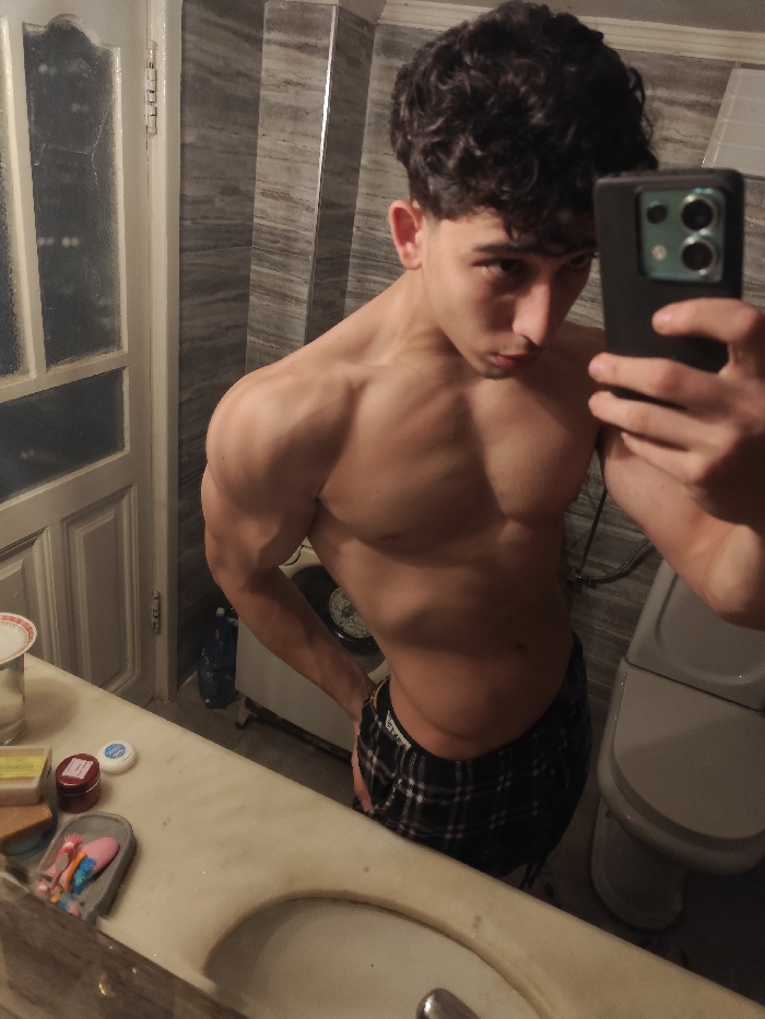 A shirtless, muscular young man with curly hair poses for a selfie in a bathroom. He wears plaid pajama bottoms. The background features a tiled wall, a partially visible washing machine, and a toilet. The foreground includes a marble countertop with toiletries and a sink. The man holds his phone in his right hand, capturing the image at a slight angle.