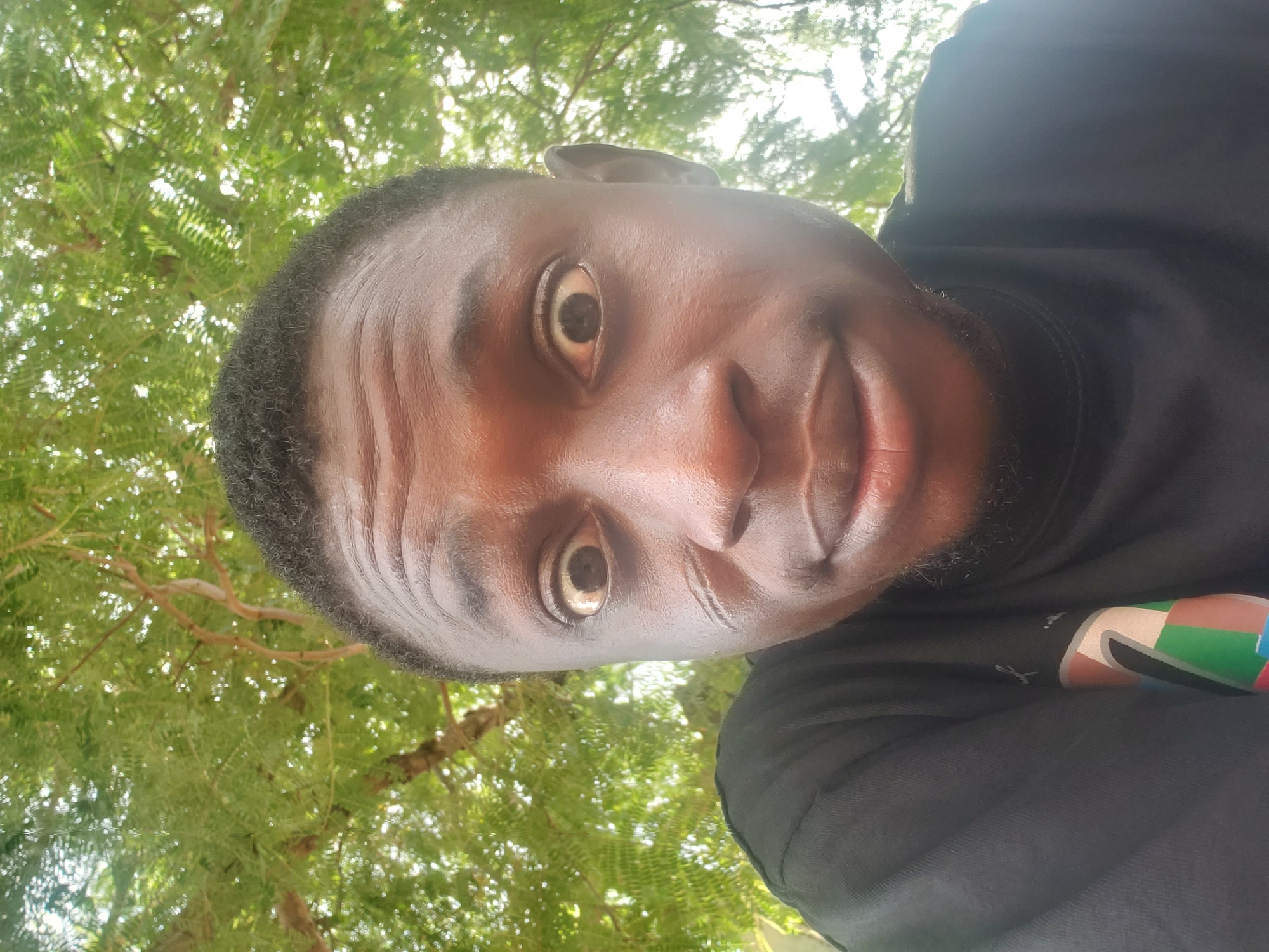 A medium-close-up shot of a Black man with short, dark hair, wearing a black shirt. The man is looking directly at the camera with wide eyes. The background shows green foliage, suggesting an outdoor setting. The image's upward angle implies the photo was taken from a low position. There is no verifiable evidence to place the photo within Adankolo, Nigeria.