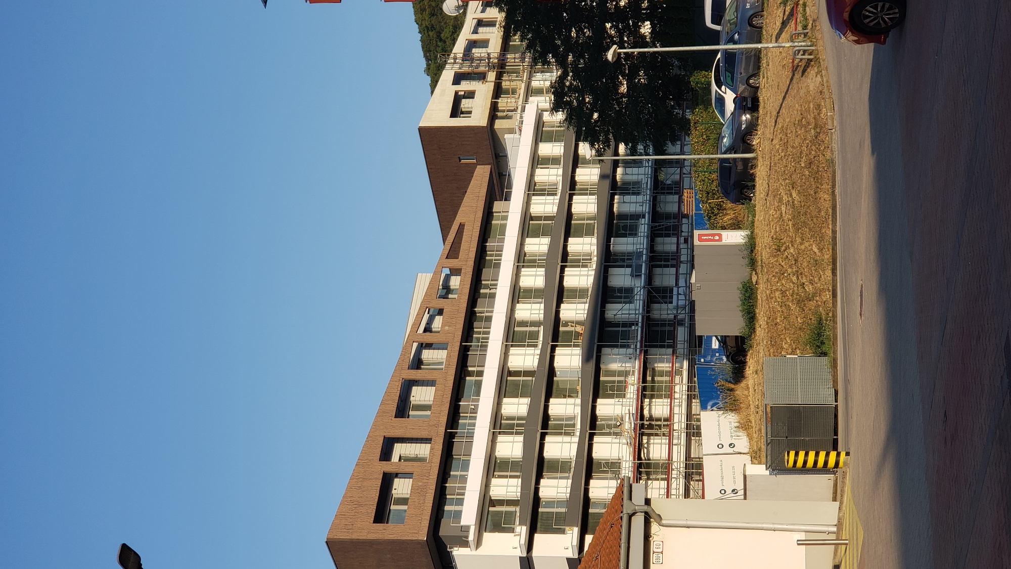 The image depicts a modern residential building under construction in Bratislava, Slovakia, likely in the Nové Mesto district, given the architectural style and urban context. The building features a distinctive, undulating facade with a brown, textured upper level and white, multi-story sections with numerous windows. Scaffolding is visible on the lower floors, indicating ongoing construction work.
In the foreground, a paved area with red brick paving is visible, leading to a grassy embankment. Several parked cars are present on the embankment, including a dark SUV, a white hatchback, and a silver hatchback. A smaller, white building with a tiled roof and a sign displaying "E965 60" is located to the left of the main building. To the right, a street lamp and a mature tree are present. The sky is clear and blue, suggesting a sunny day.
There are no people or animals visible in the image. The overall impression is one of urban development and progress, with a focus on modern architecture and infrastructure.